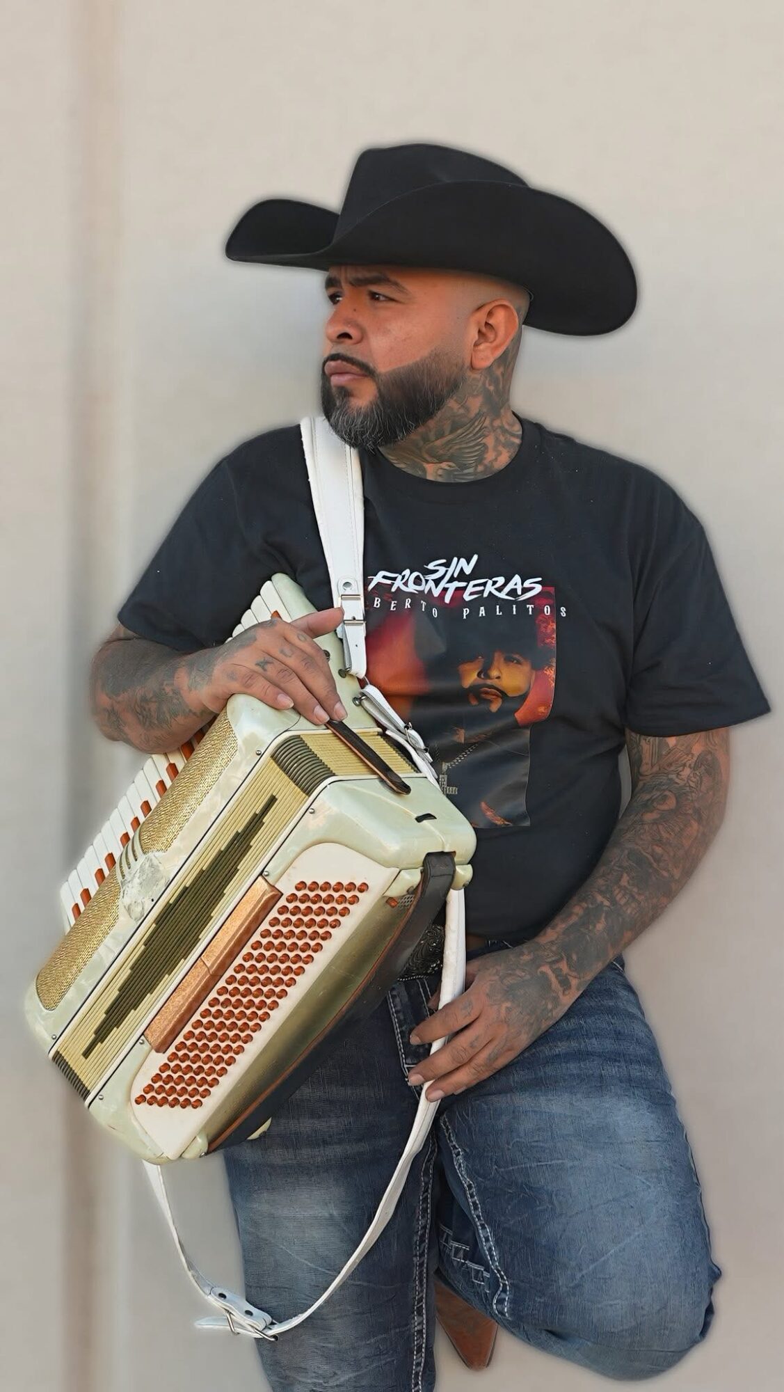 Man with a beard and tattoos wearing a black cowboy hat and T-shirt, holding a vintage radio, standing against a plain wall.