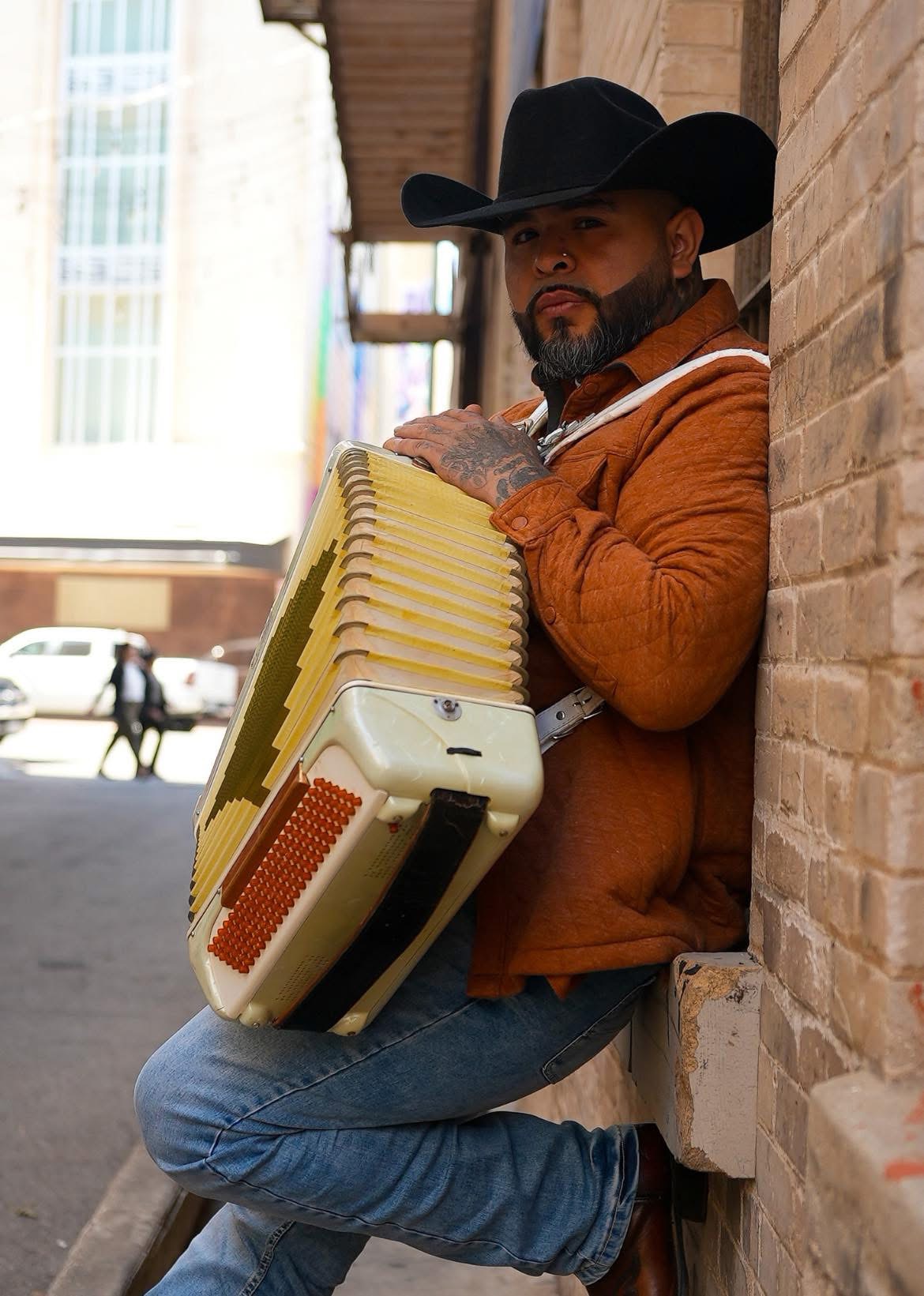 Man with a beard wearing a black cowboy hat and orange jacket leaning against a brick wall, holding an accordion.
