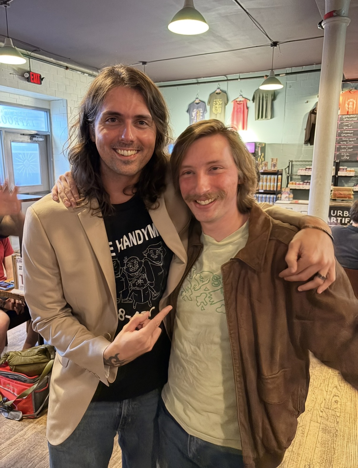 Two smiling men with shoulder-length hair, one in a beige blazer and the other in a brown jacket, pose indoors.