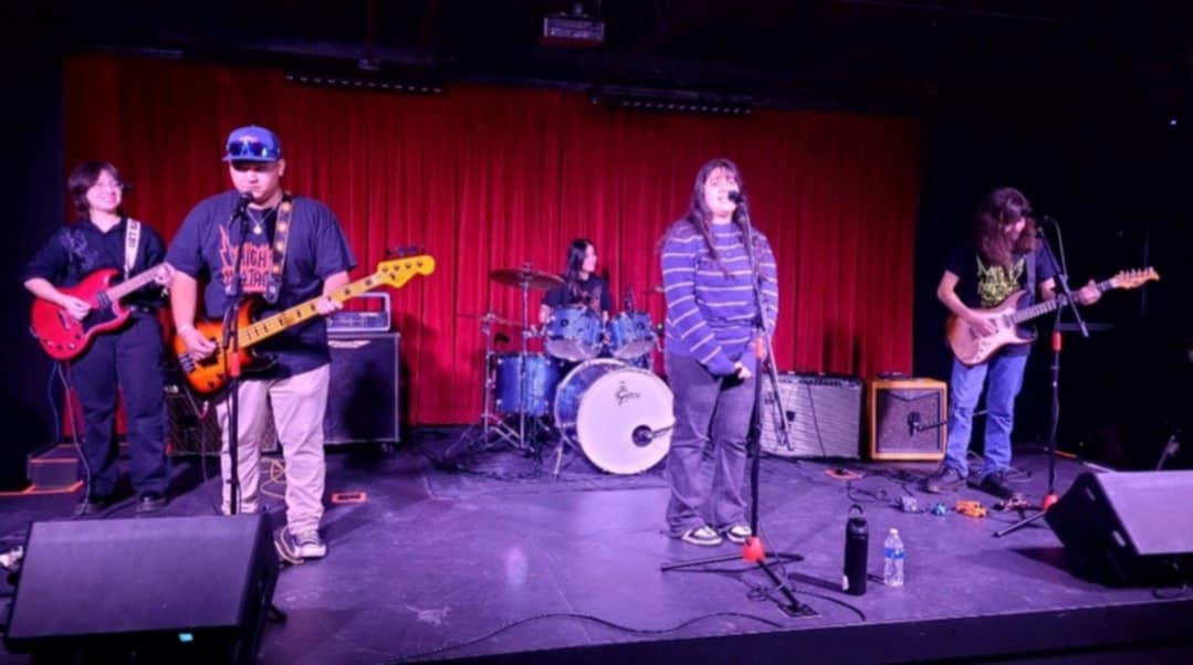 Band performing on stage with red curtain background, four members playing guitars, drums, and singing, stage equipment visible.
