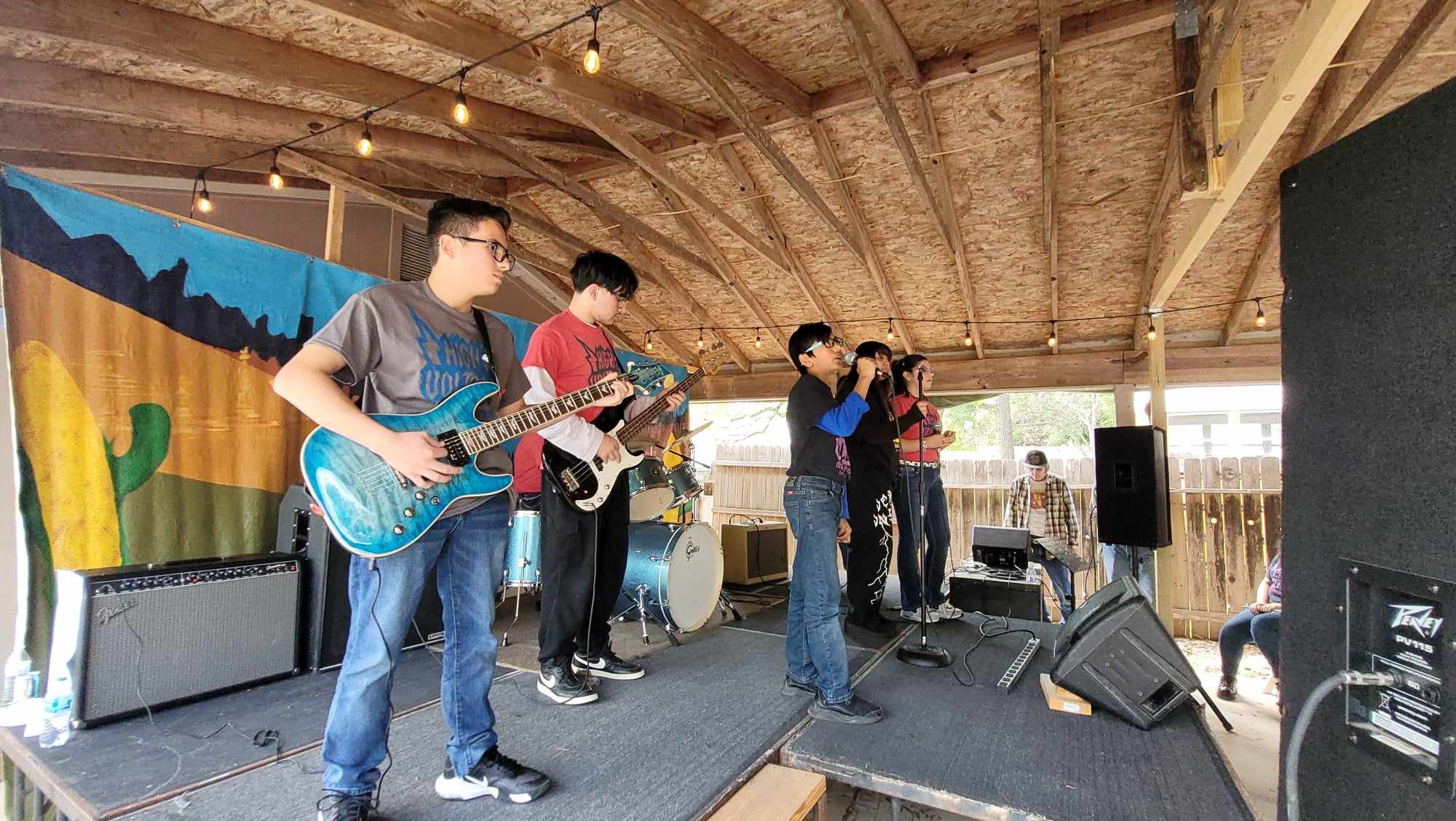 Group of young people performing music on a stage with instruments, under a wooden ceiling, with a colorful backdrop and speakers.