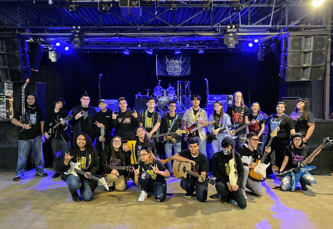 Group of people with guitars on stage, some standing and some kneeling, in a music venue with blue lighting.