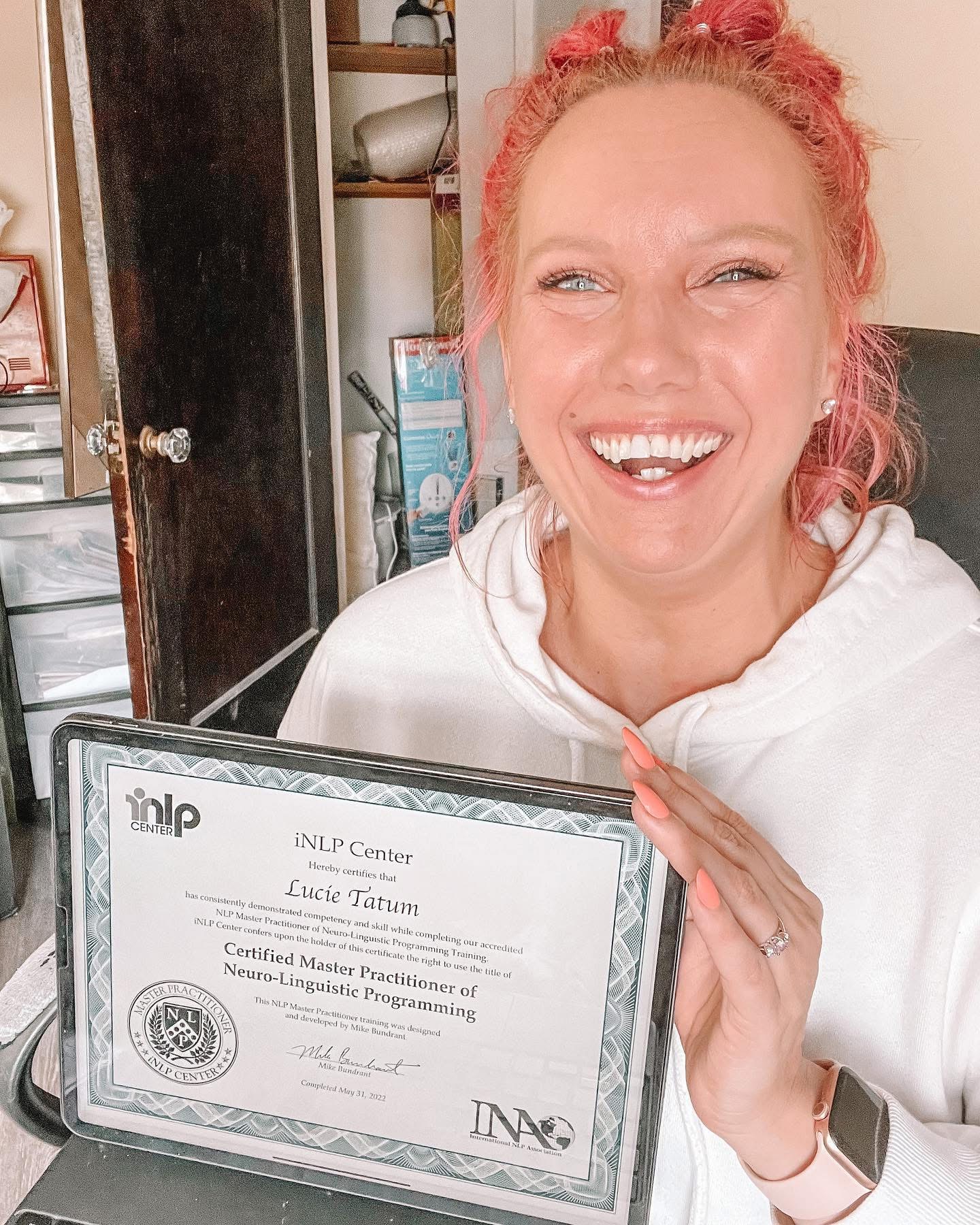 Woman with pink hair smiling, holding a certificate, in a room with shelves and a door behind her.