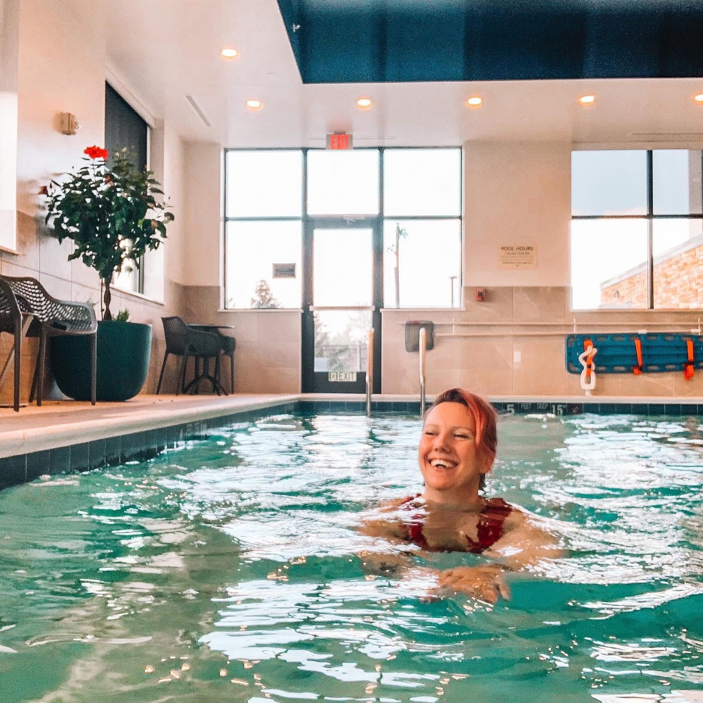 Woman smiling and swimming in an indoor pool with large windows and plants, bright natural light, and pool safety equipment visible.