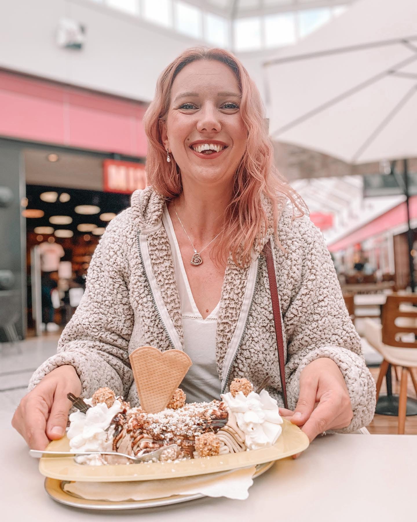 Smiling woman with pink hair holding a plate of dessert at an outdoor cafe, umbrella overhead, background of shops and seating.