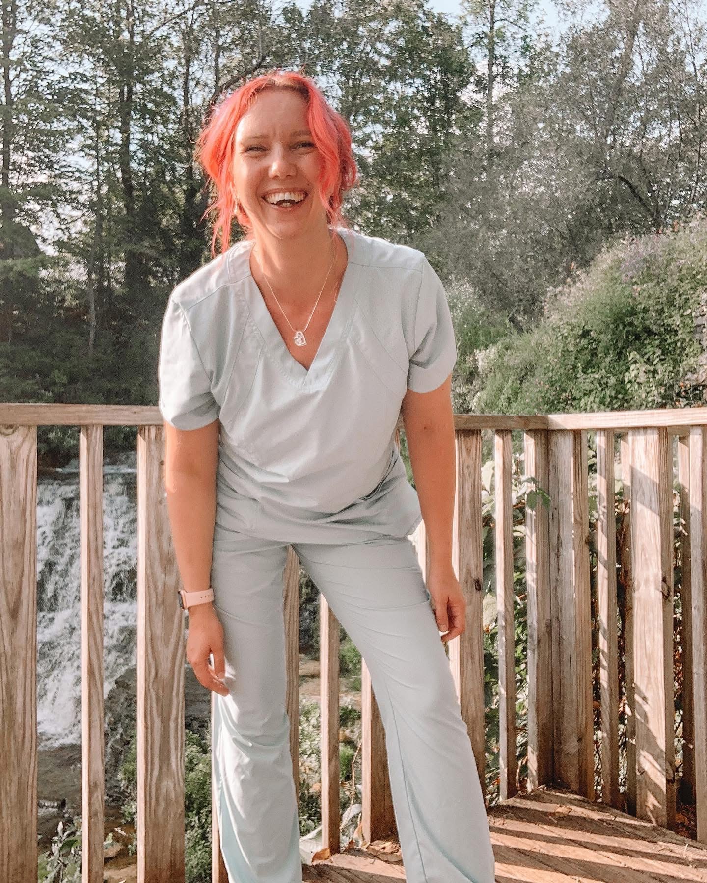 Woman with pink hair smiling outdoors on a wooden deck, trees and bushes in background.