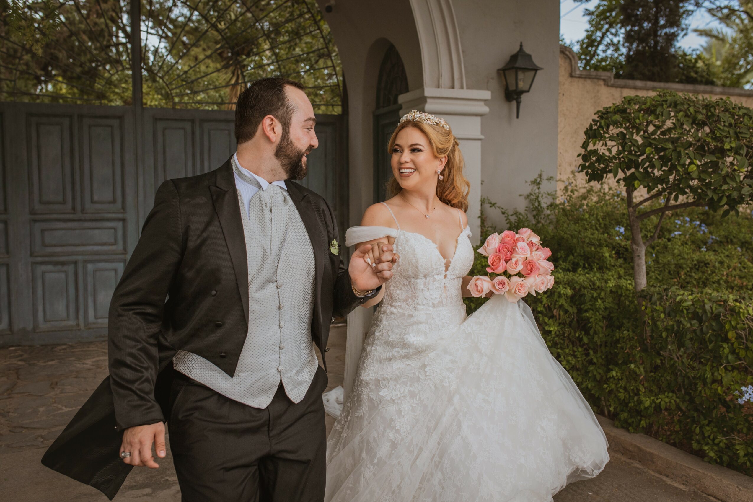 A bride and groom walk outdoors, smiling, with the bride holding a bouquet of pink flowers, in front of a gate and greenery.
