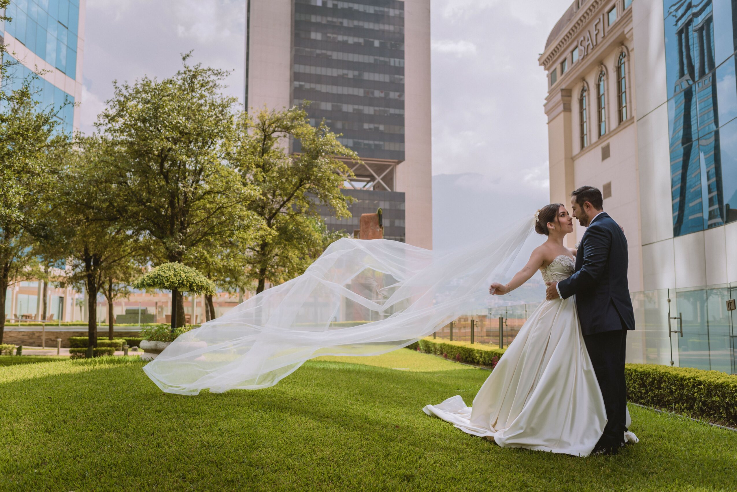 A bride and groom stand on grass in an urban park, with the bride's veil flowing in the wind, surrounded by modern buildings.