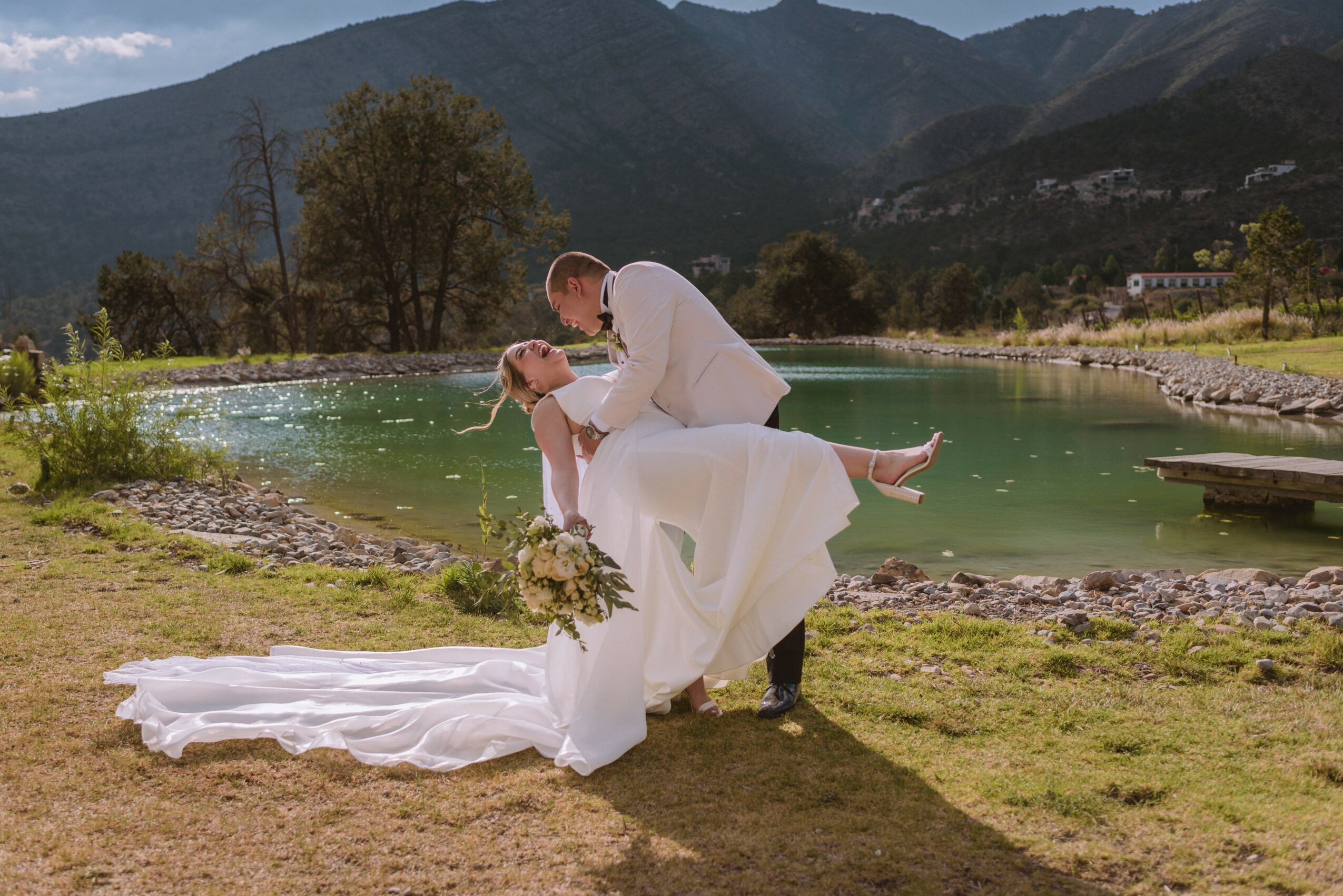 A couple in wedding attire by a lake with mountains in the background, the groom holding the bride who is leaning back.