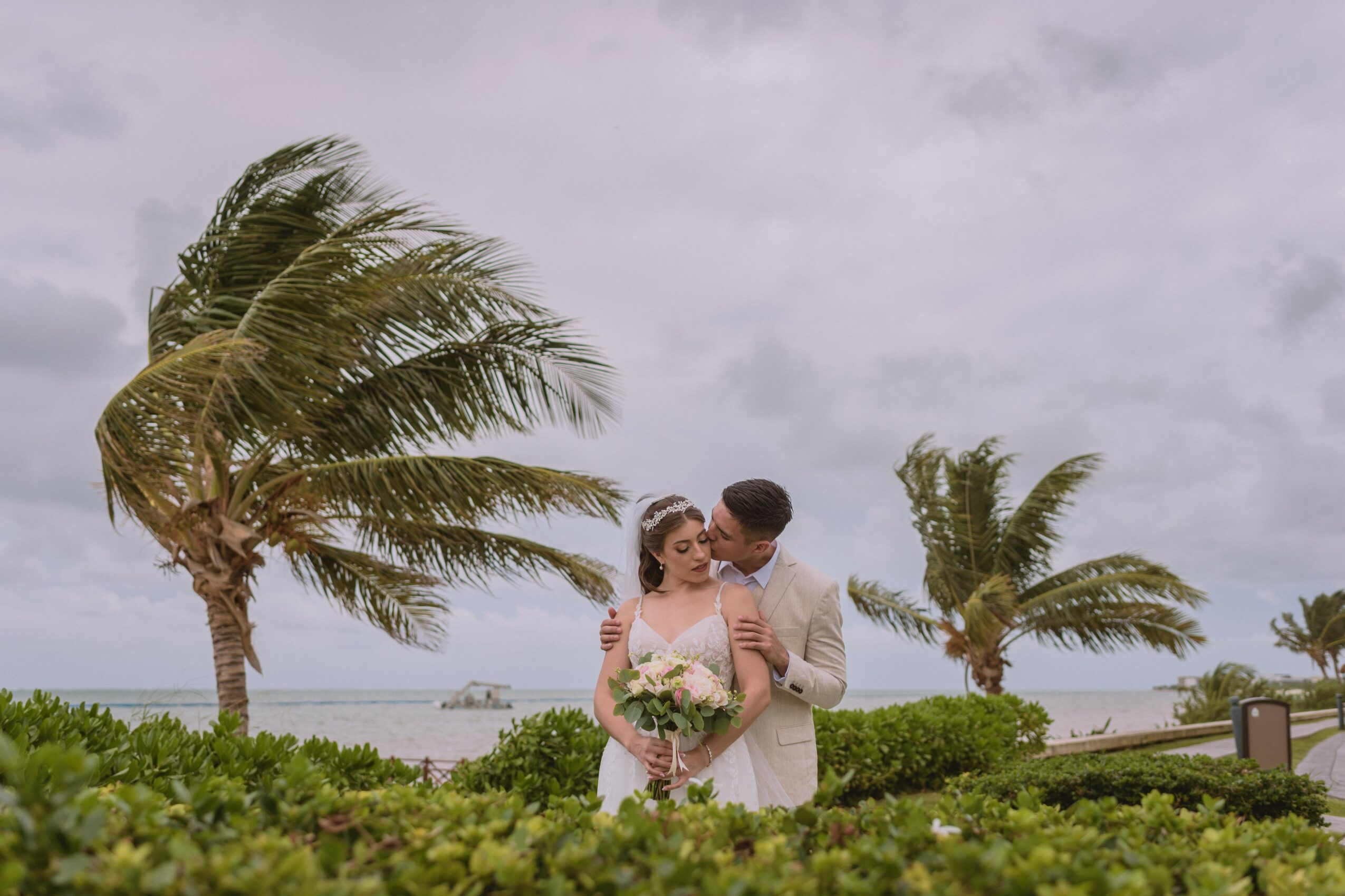 Couple standing outdoors with palm trees and cloudy sky, holding a bouquet of flowers, embracing each other.