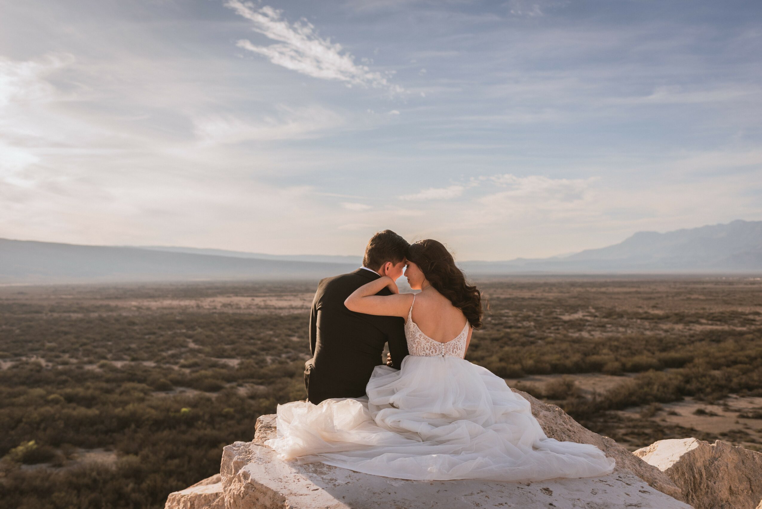 A couple sitting on rocks outdoors, embracing, with a landscape and sky in the background.