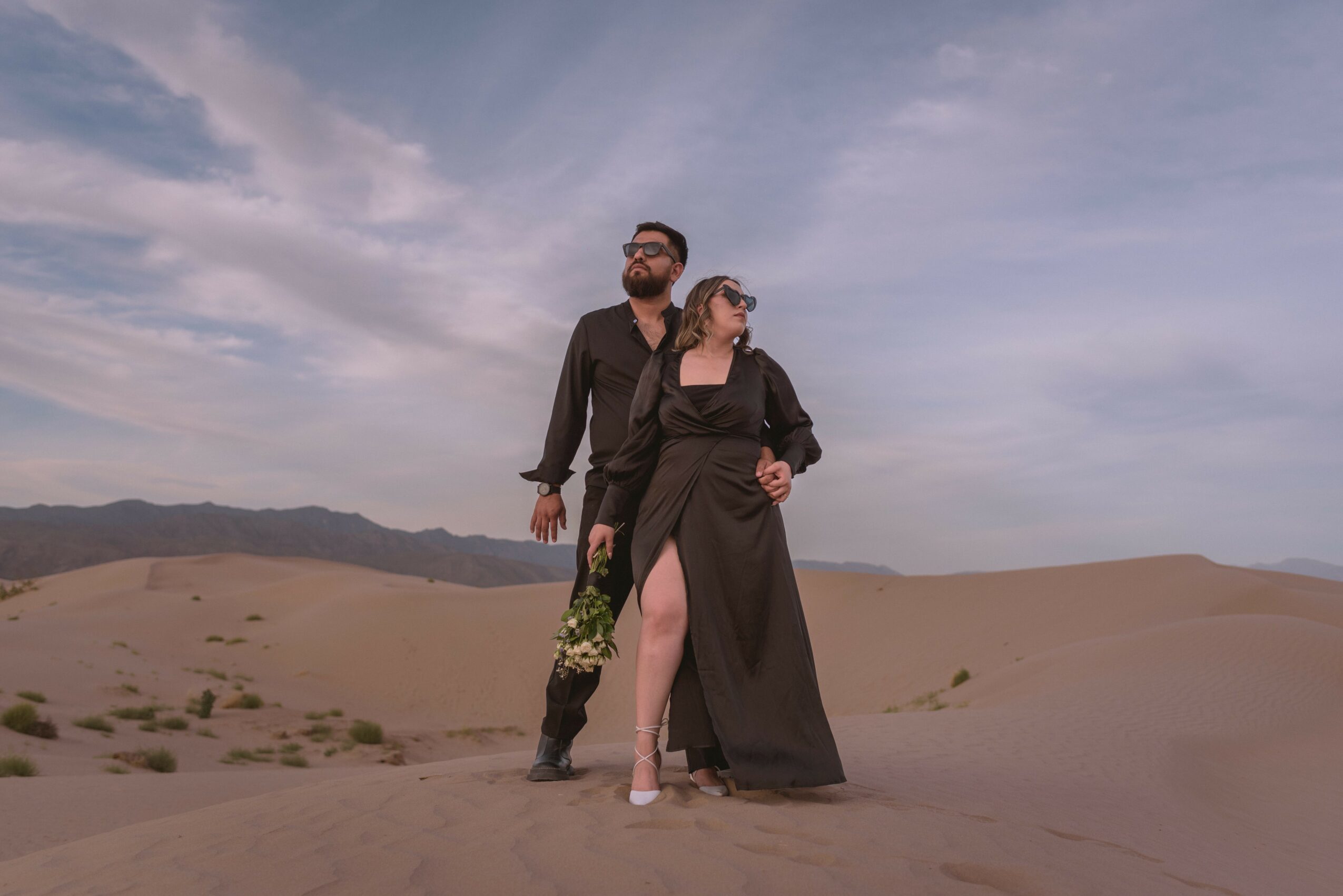 A man and woman stand in a desert landscape with sand dunes and a cloudy sky, dressed in dark clothing, looking into the distance.