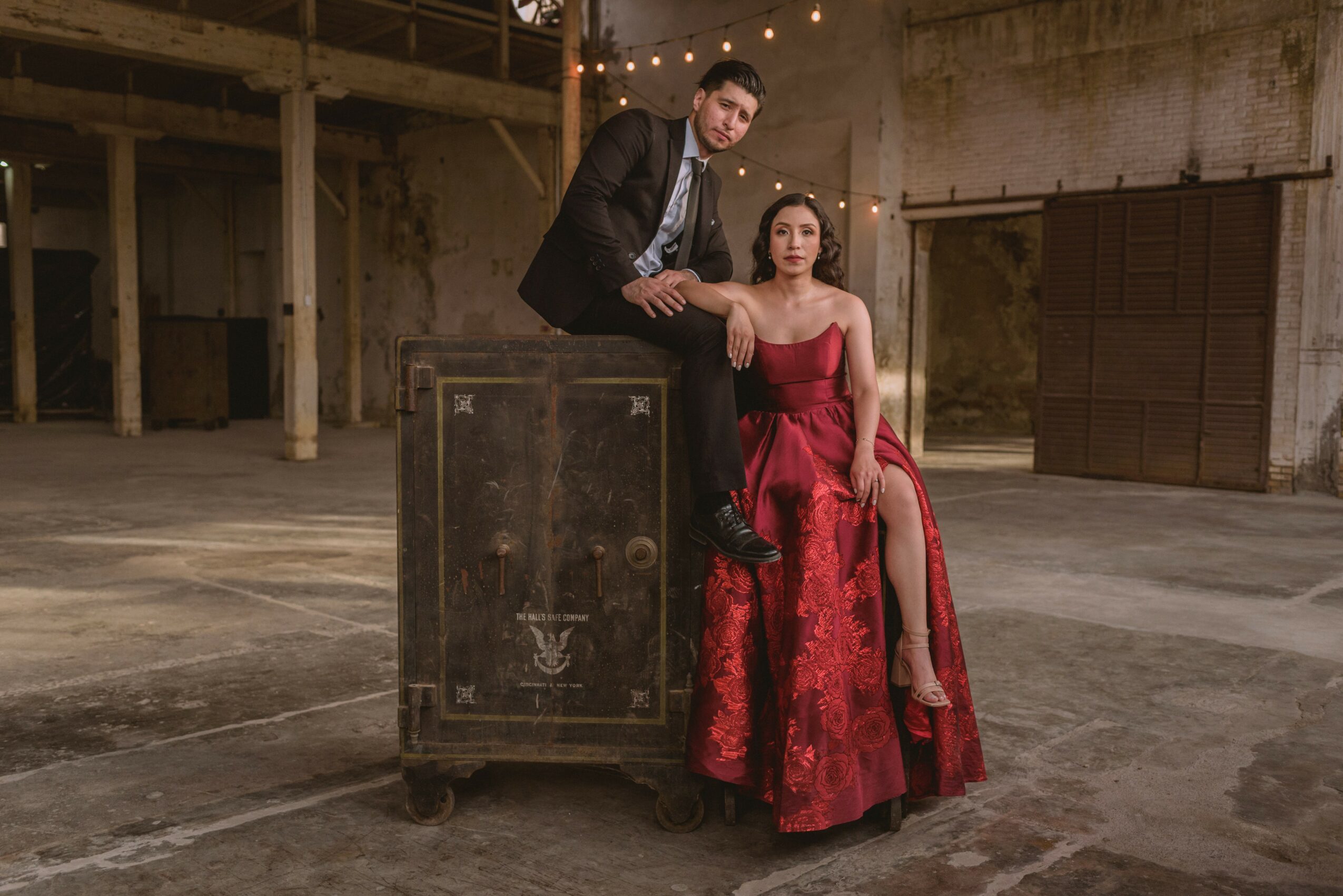 A man in a suit and a woman in a red gown pose in an industrial setting with string lights overhead.