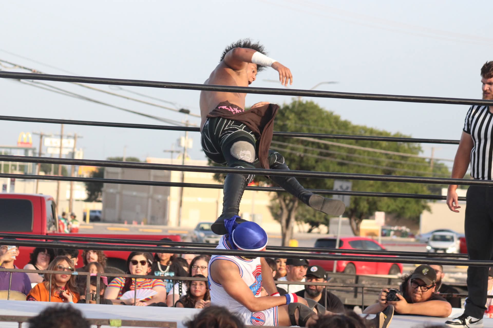 Wrestler jumps onto opponent in ring with referee and audience watching outdoors.
