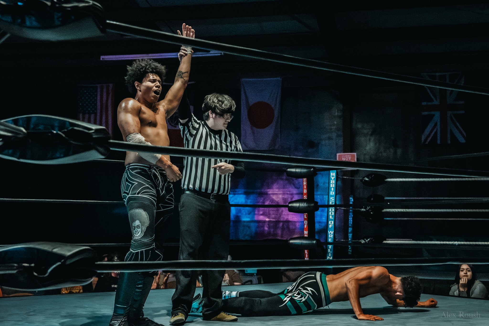 A wrestler with curly hair raises a sword, a referee stands nearby, and another wrestler lies on the mat in a wrestling ring.