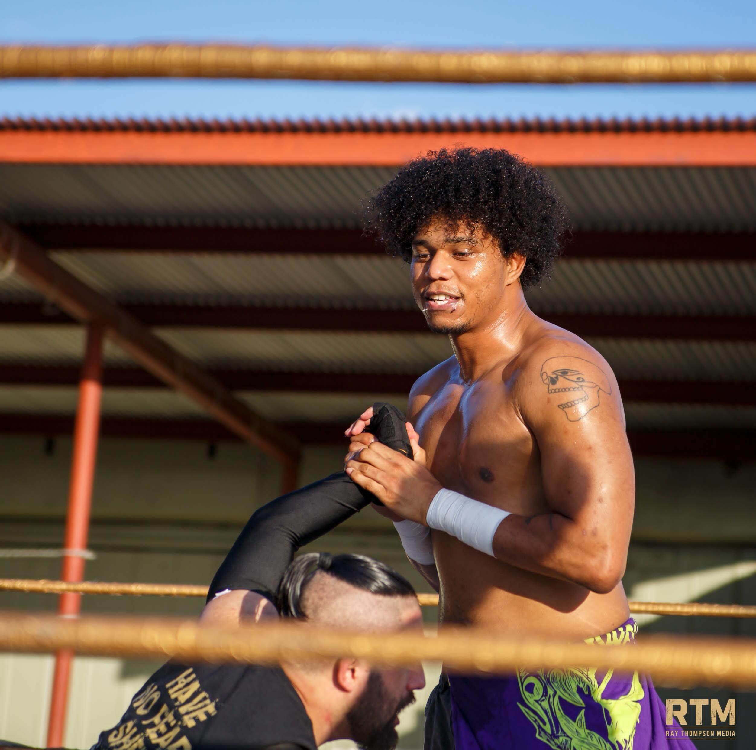 Boxer with curly hair and tattoos, holding a towel, standing in a boxing ring outdoors.
