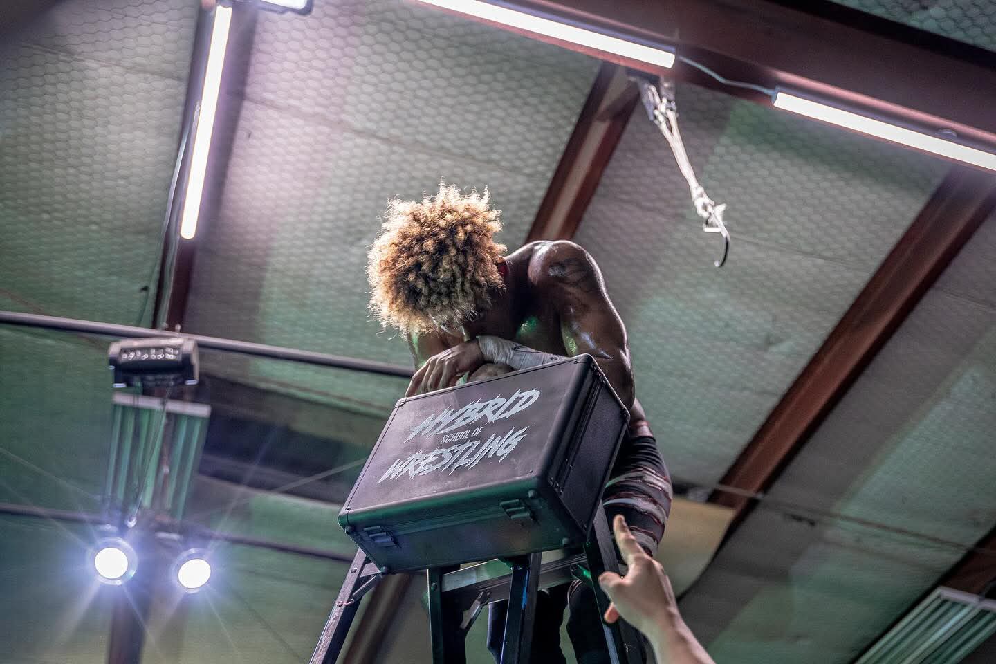 Two people embrace in a warehouse or industrial space with a high ceiling and metal beams, one holding a television set.