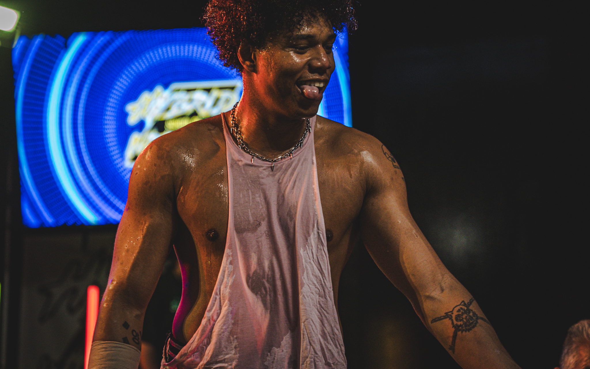 Person with curly hair smiling, wearing a wet sleeveless shirt, in front of a blue circular background, possibly at a concert or event.