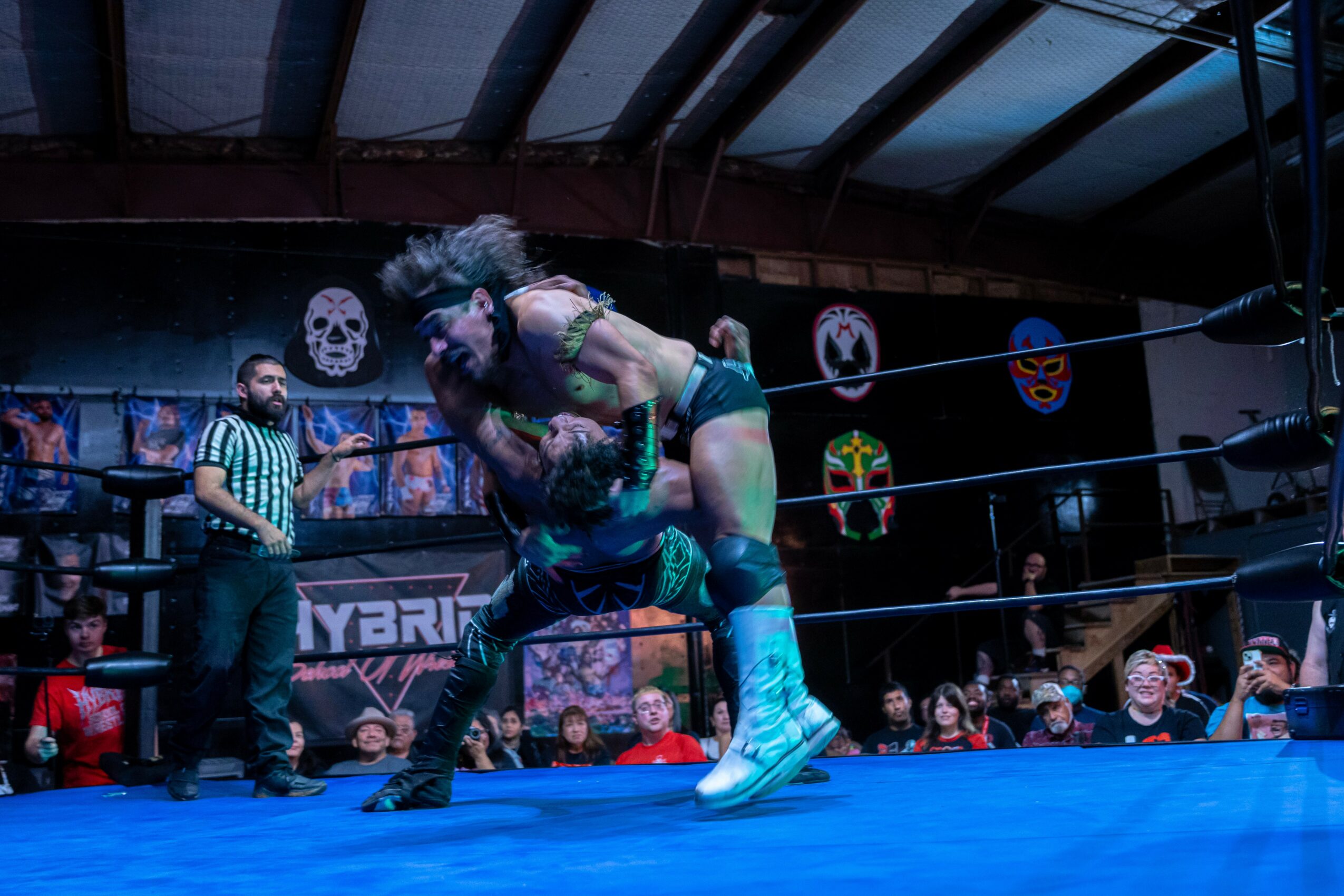 Two wrestlers engaged in a match inside a ring, with a referee and audience watching, colorful masks on the wall.