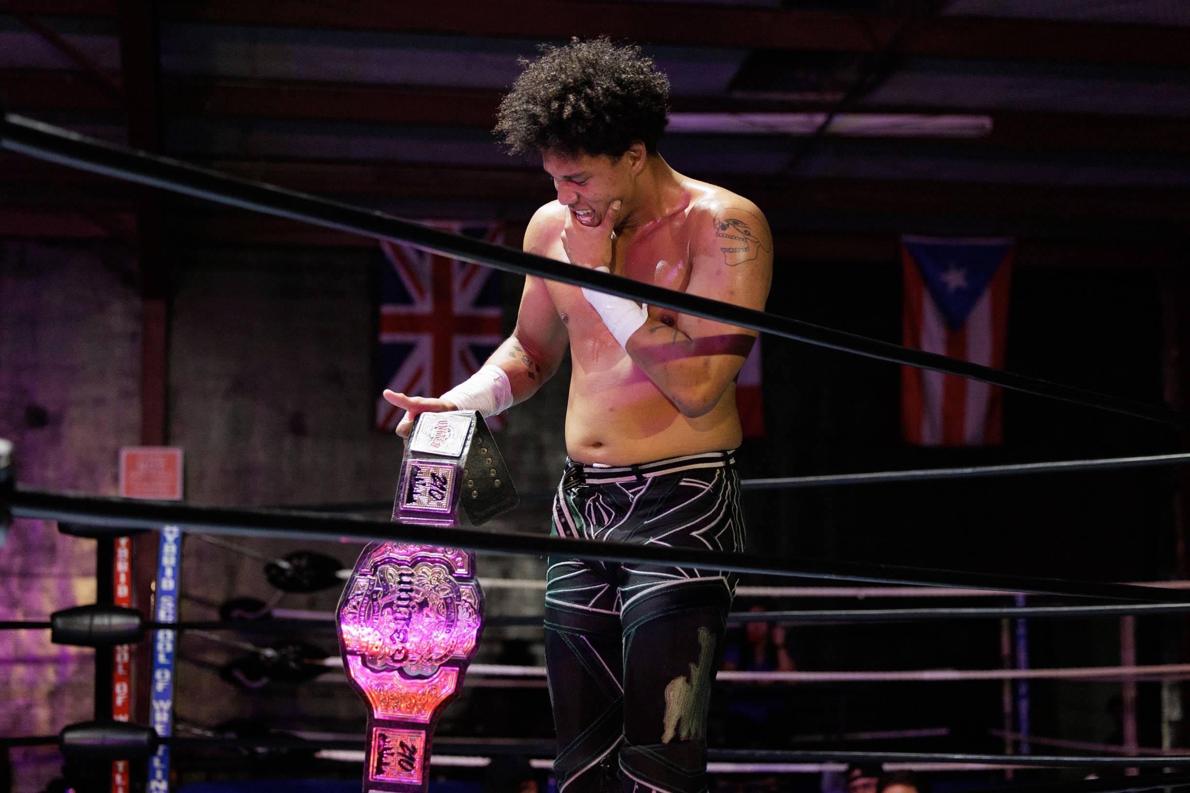 A shirtless male wrestler with curly hair stands in a wrestling ring, holding a championship belt, with flags in the background.