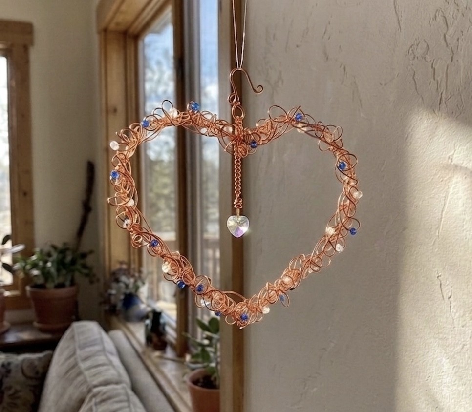 Heart-shaped wire decoration with beads hanging near a window, sunlight illuminating the wall.