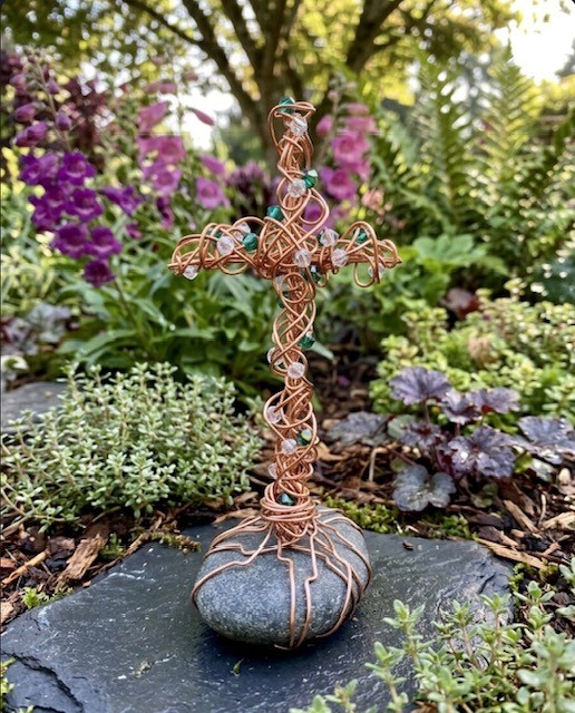 Wire cross sculpture on a rock surrounded by colorful garden flowers and greenery.