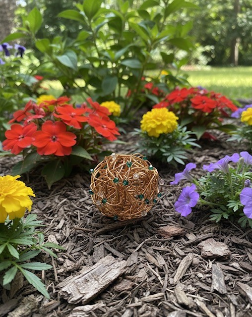 Colorful flowers including red, yellow, and purple blooms in a garden bed with mulch, greenery, and trees in the background.