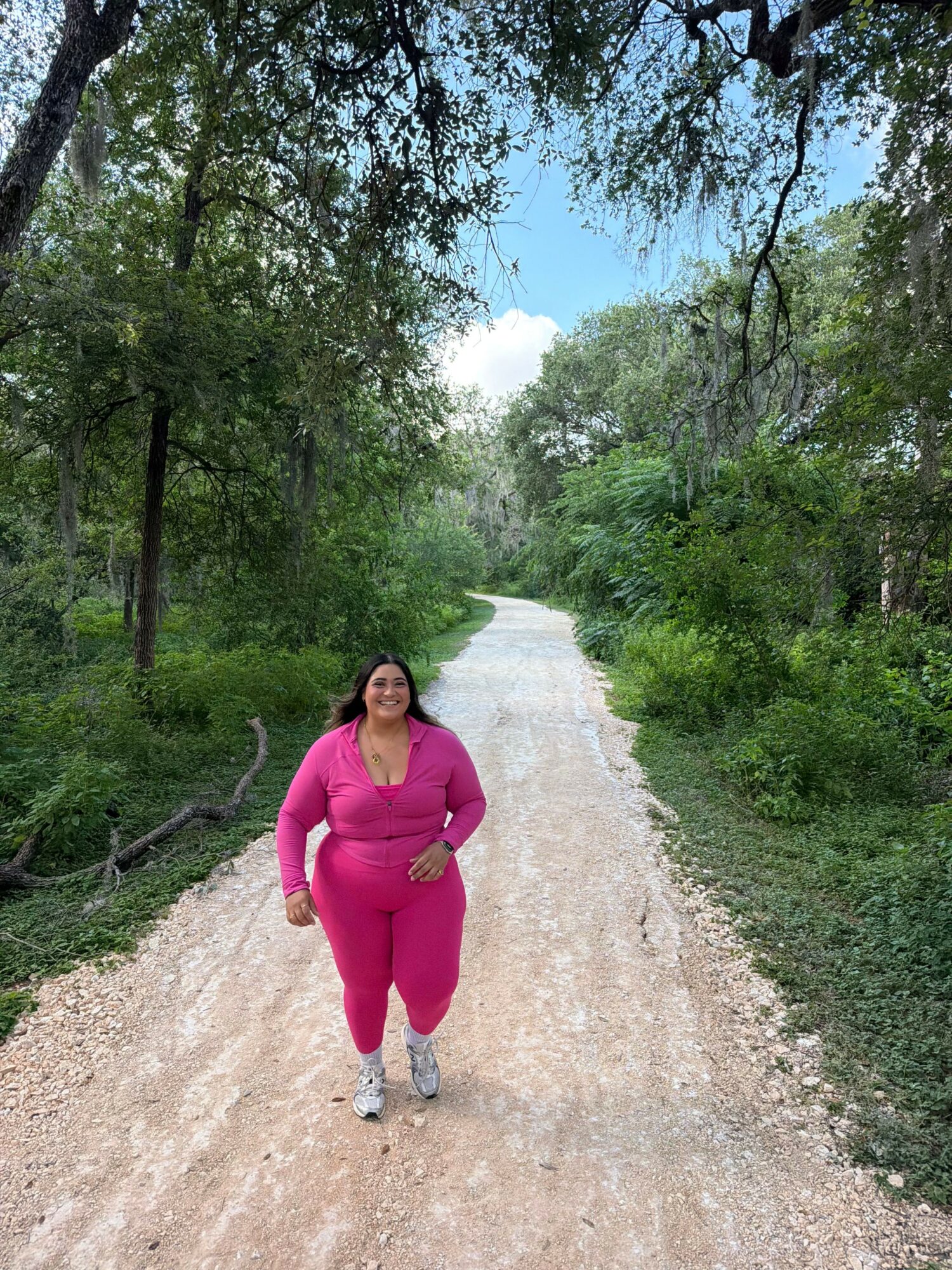 Woman in pink outfit walking on a dirt path surrounded by green trees and blue sky.