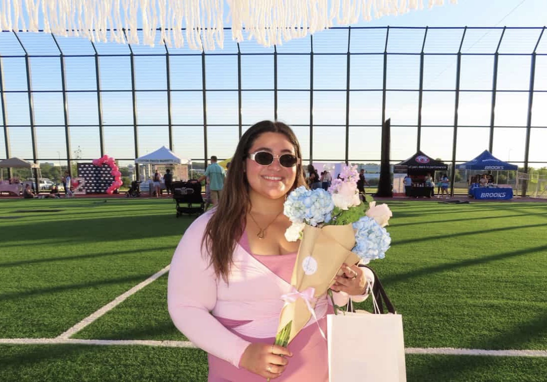 Woman holding flowers and a shopping bag, smiling outdoors on a sunny day, with a sports field and tents in background.