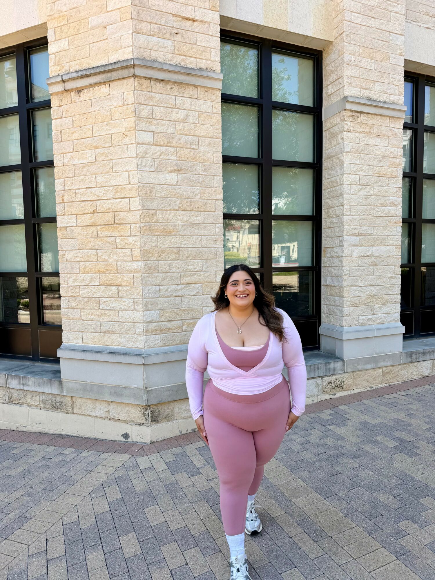 Smiling woman in pink and white outfit walking on sidewalk in front of a brick building with large windows.