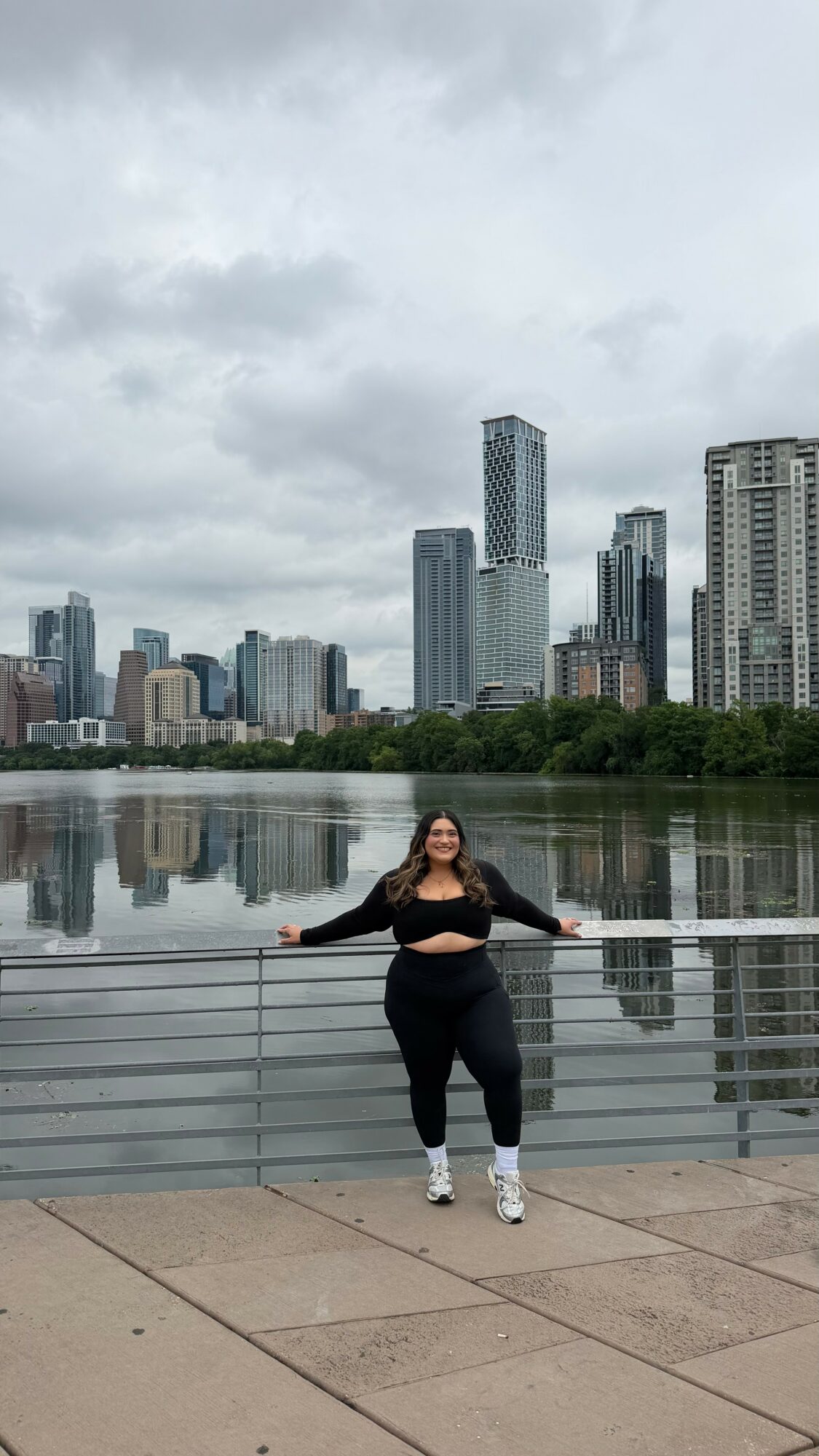 Person standing by a railing with city skyline and river in background under cloudy sky.