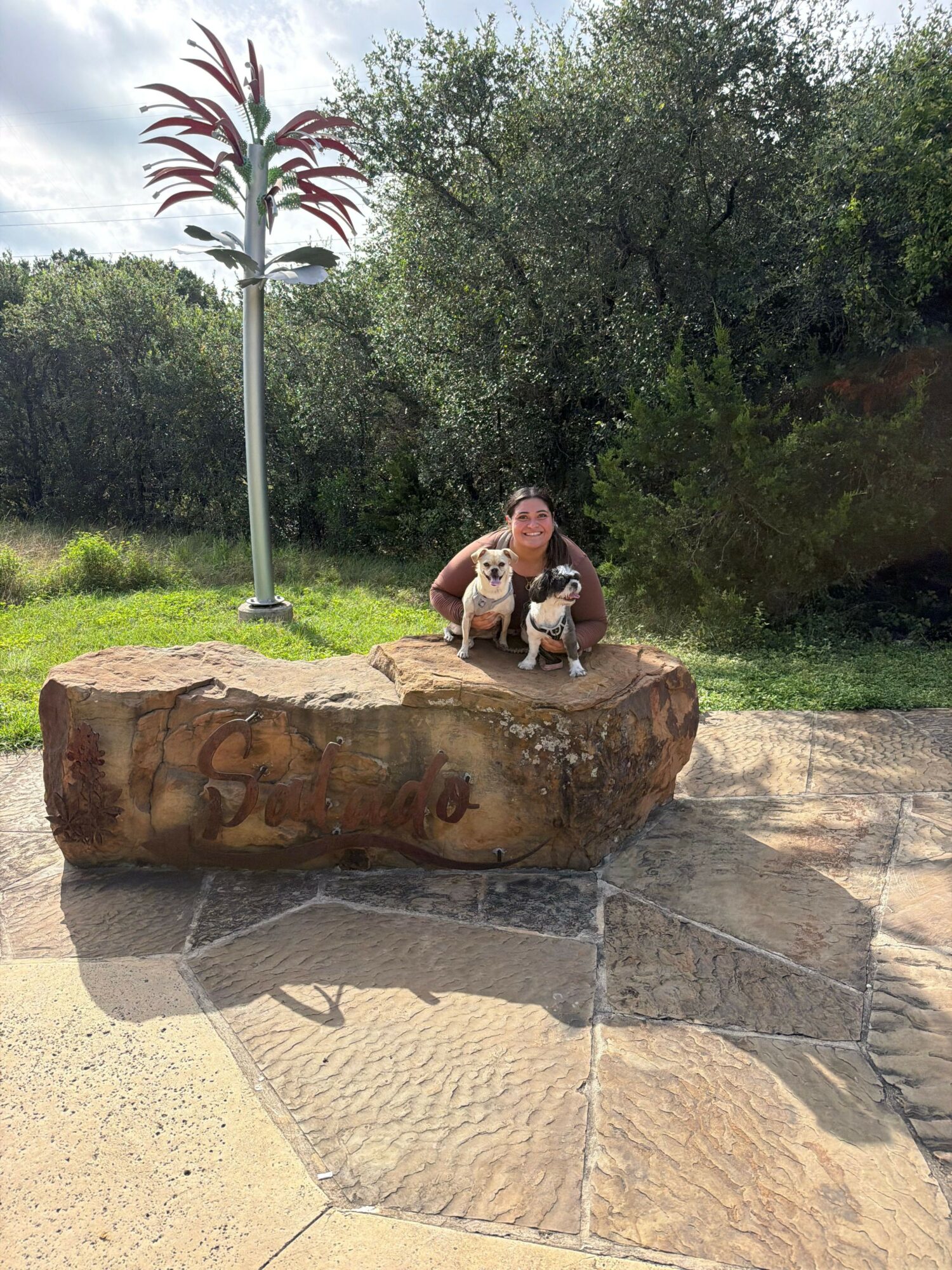 Two people sit on a large, rounded stone in a park with trees and grass, taking a selfie.