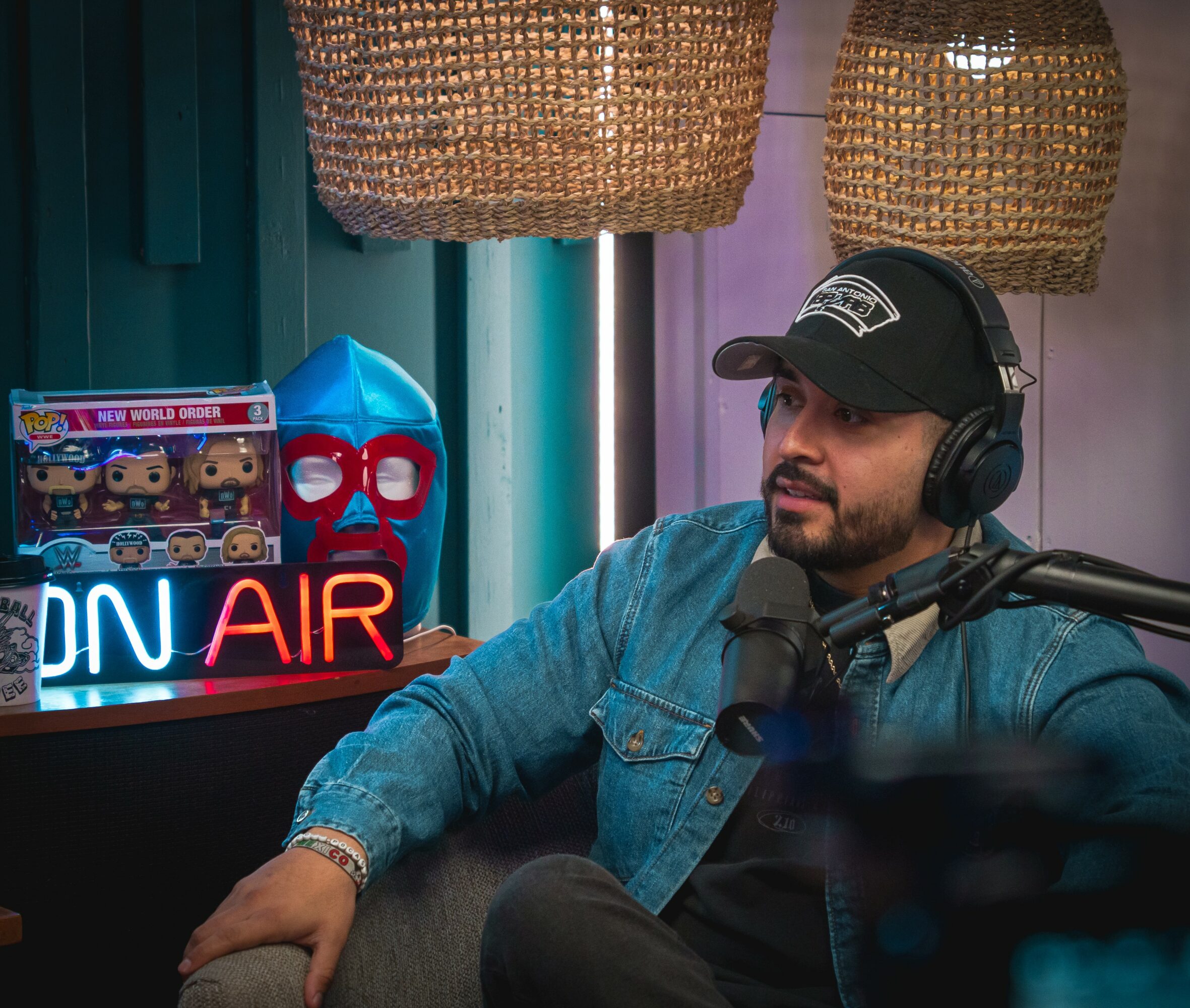 Man wearing headphones and a cap, sitting in a podcast studio with decorative baskets and a neon sign.