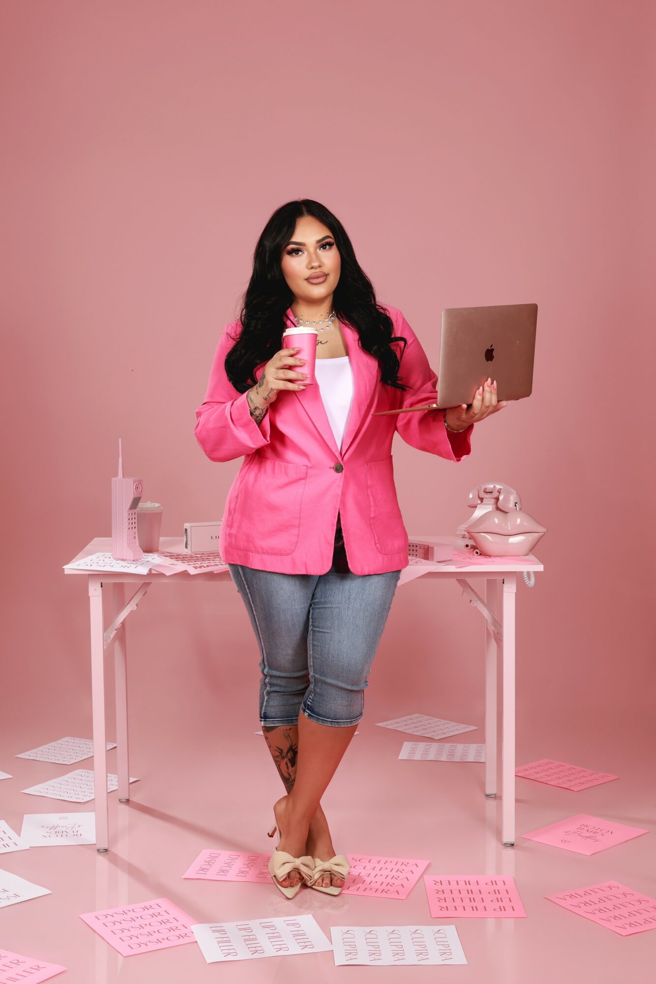 Woman in pink blazer holding a coffee cup and laptop, standing at a desk with office supplies, pink background.