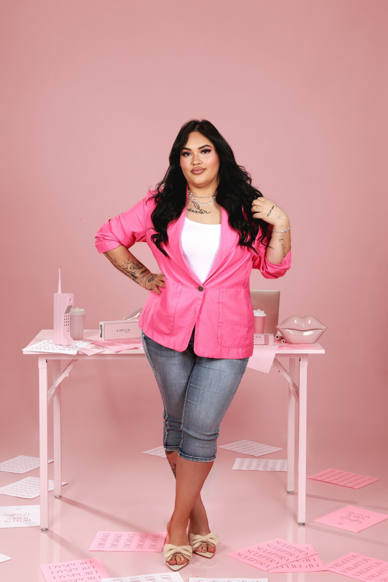 Woman with long dark hair in pink blazer and jeans standing in front of a desk with papers and office supplies, pink background.