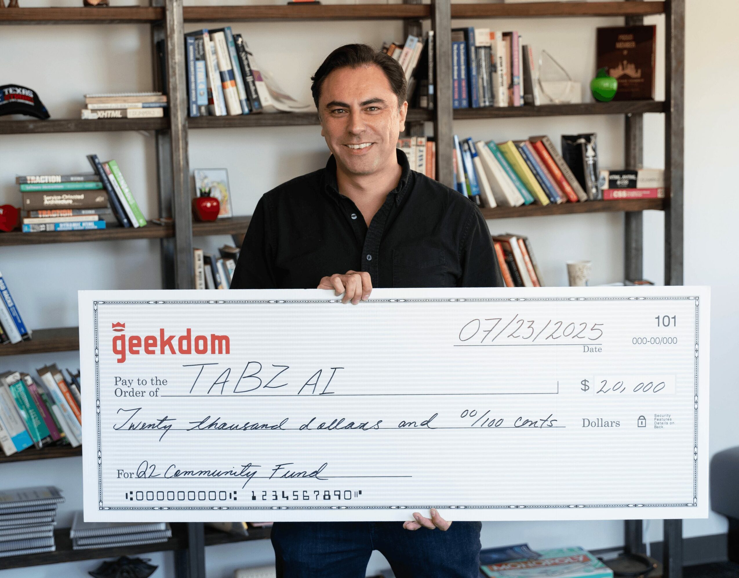 Man holding a large check in front of bookshelves, smiling, check made out to TABZ AI, dated 07/23/2025.
