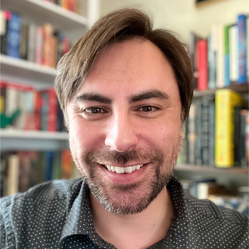 Man with brown hair and beard smiling in front of a bookshelf filled with books