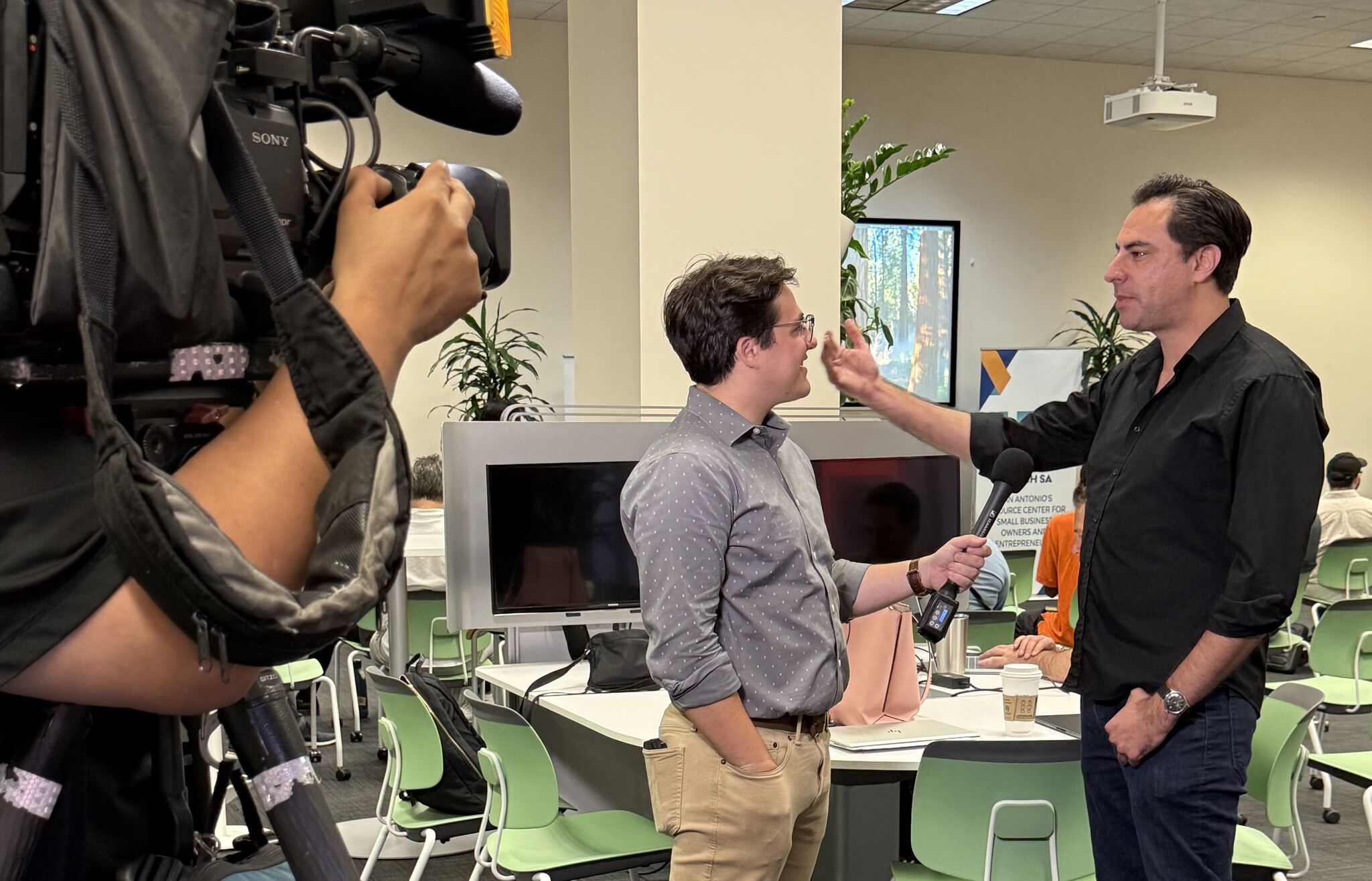 Two men are in a conversation in an office, one holding a microphone, with a camera recording them. Office has computers and green chairs.