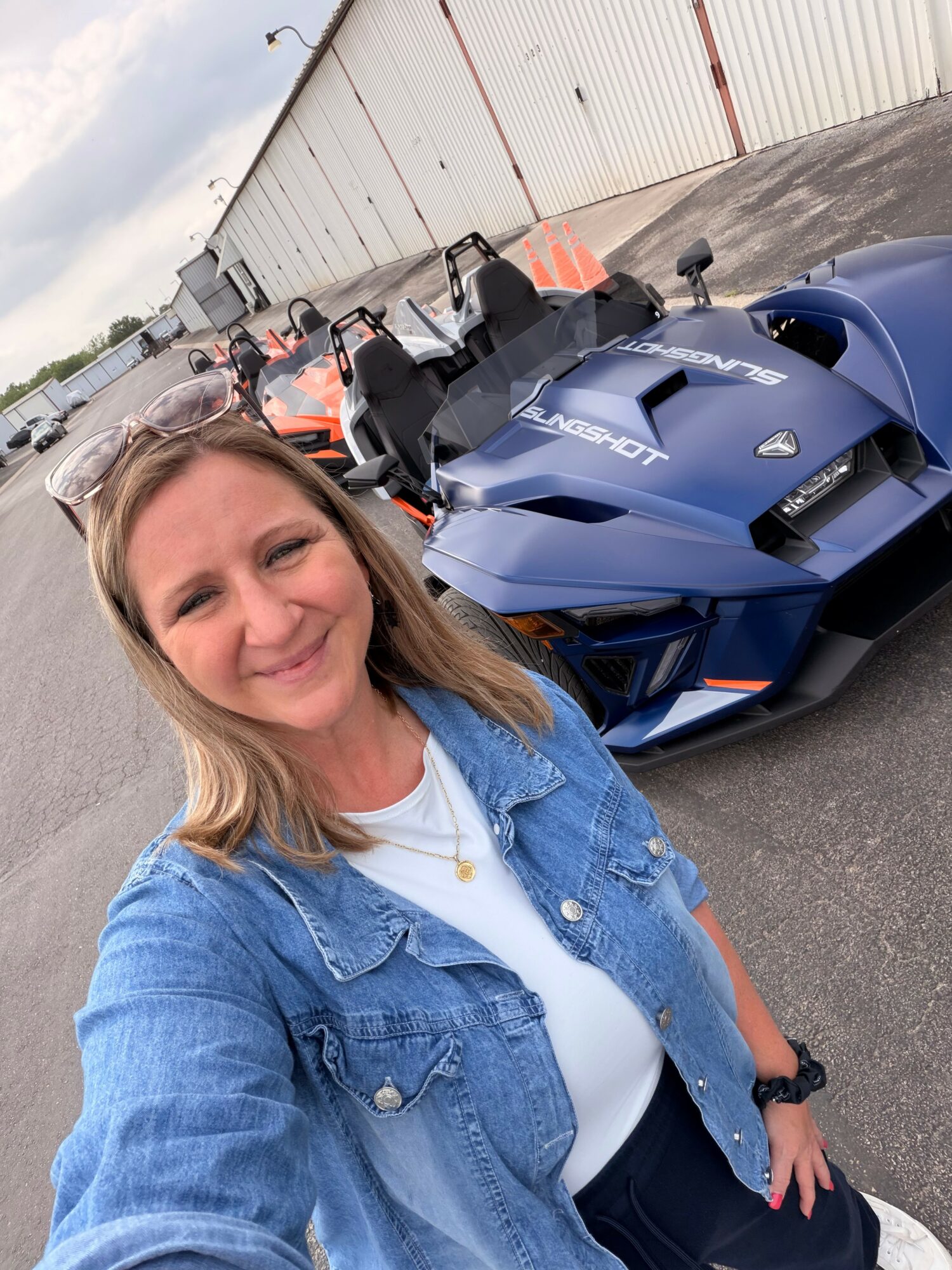 Woman taking selfie with a blue Polaris Slingshot vehicle and other vehicles in the background.