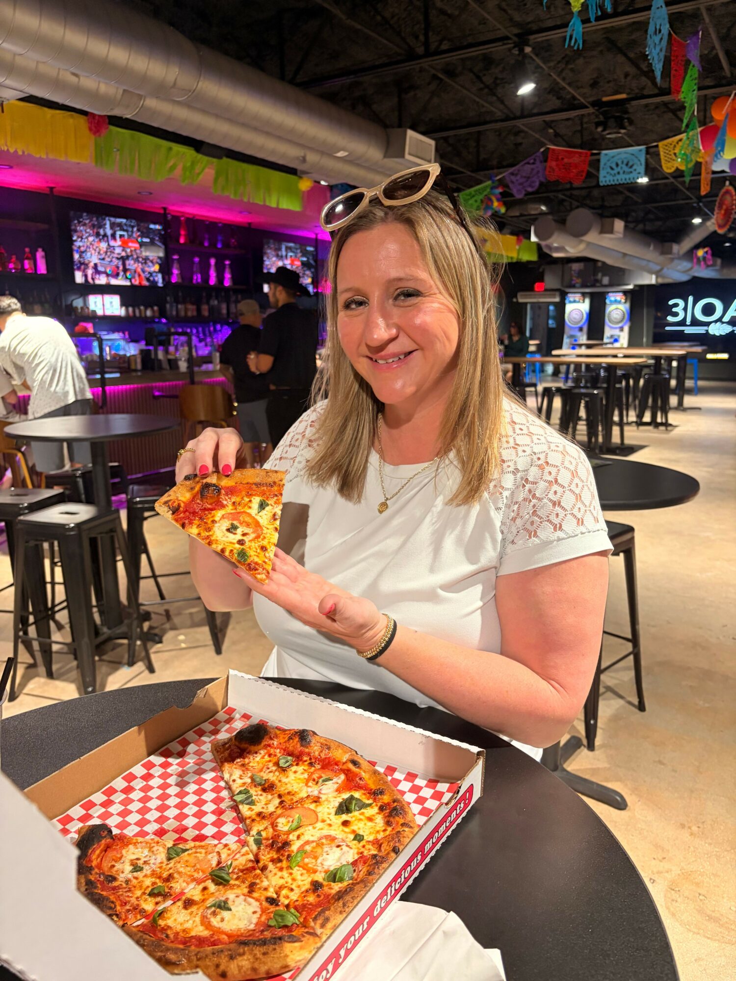 Woman sitting at a table holding a slice of pizza, with pizza boxes on the table, in a restaurant setting.