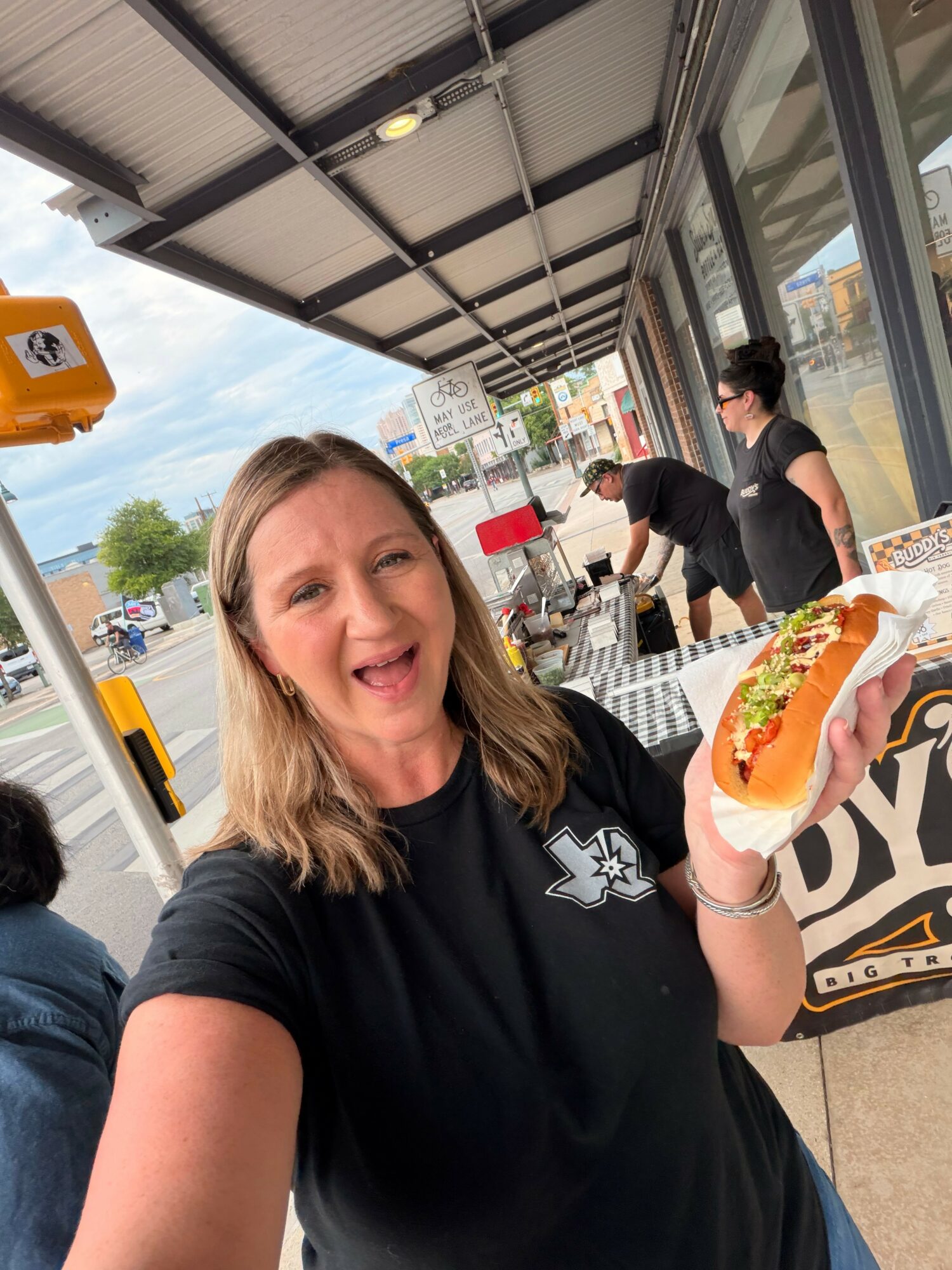 Woman smiling and holding a hot dog outside a restaurant, with other people and a parking lot in the background.