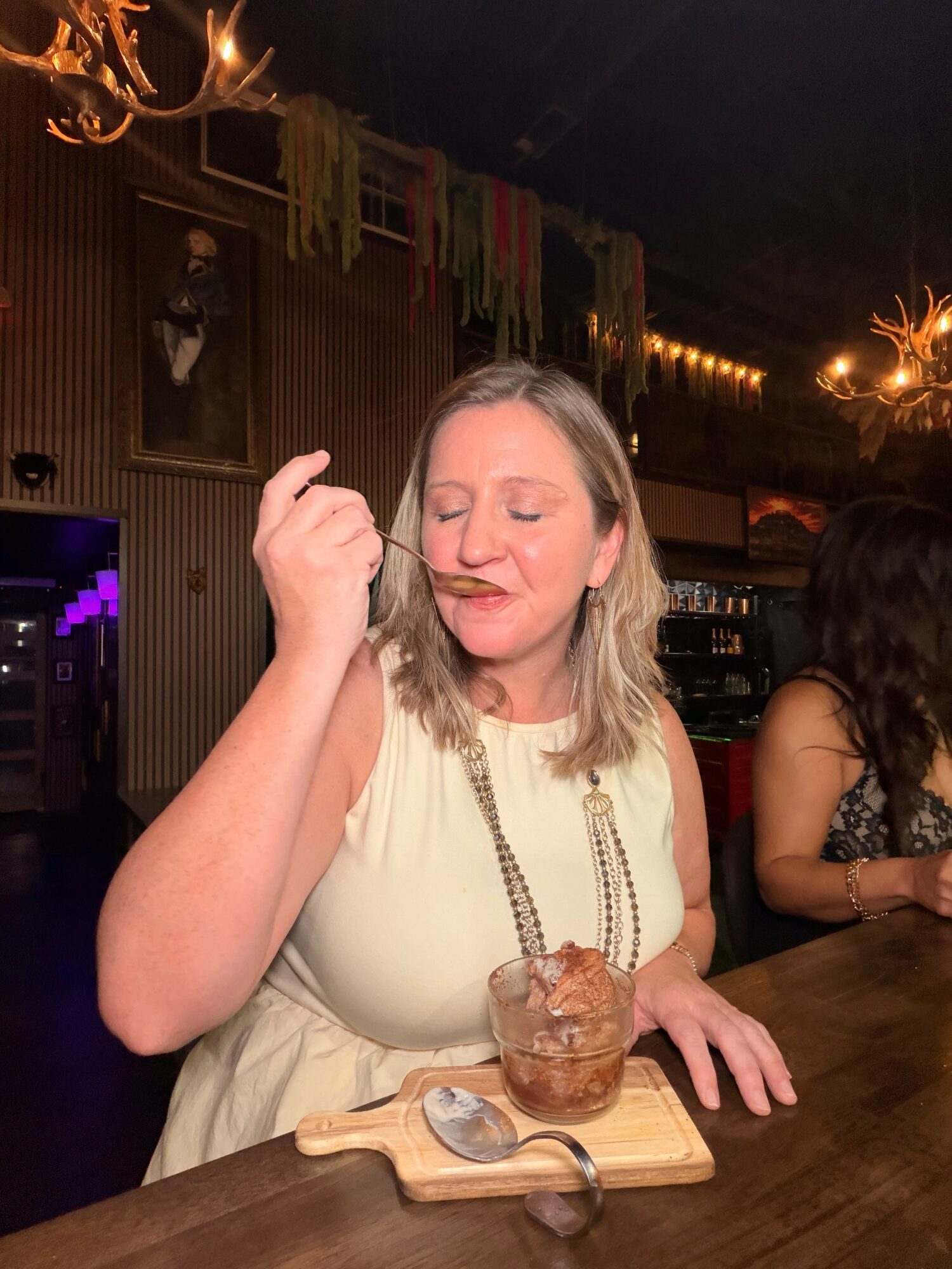 Woman with blonde hair in a sleeveless dress eating dessert at a bar, with other people and warm lighting in background.