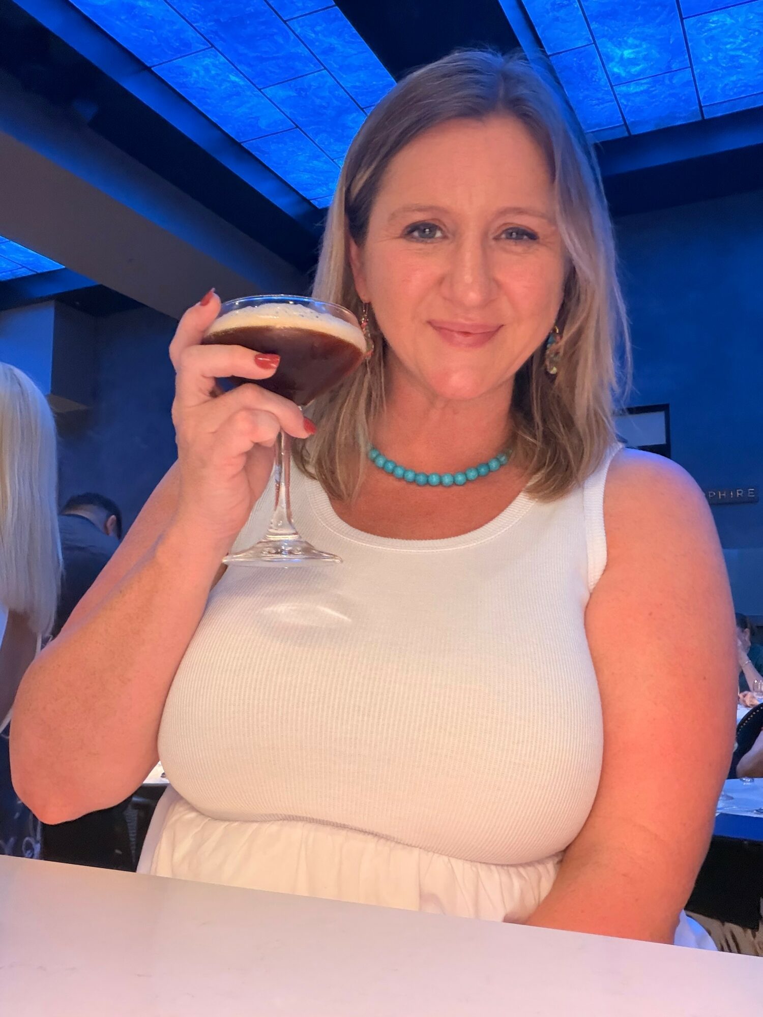 Woman with shoulder-length hair holding a cocktail glass, smiling, in a dimly lit setting with blue ceiling lights.