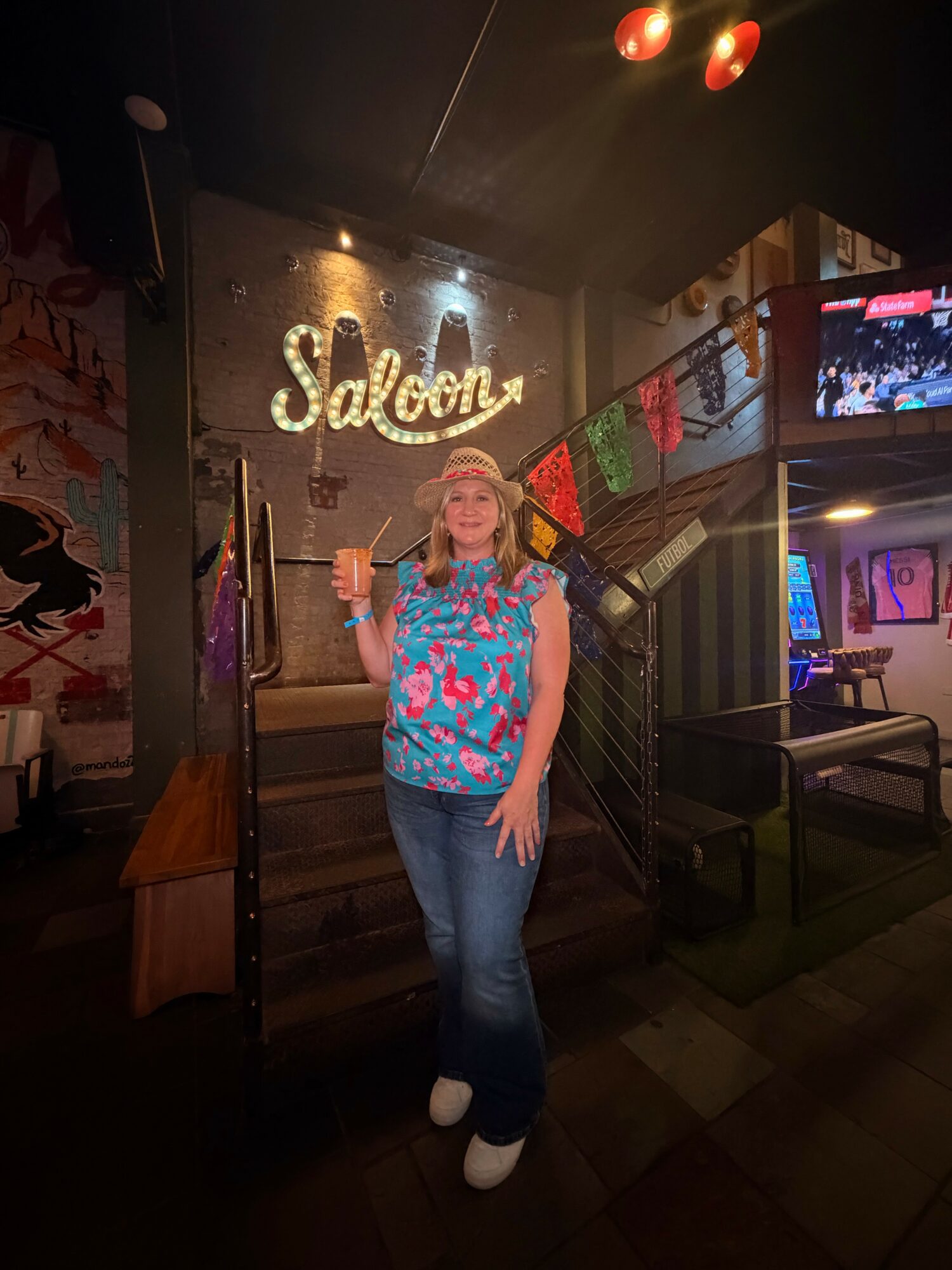 Woman standing in front of a neon sign that says 'Saloon' inside a bar or club, holding a drink, smiling, with colorful decorations around.