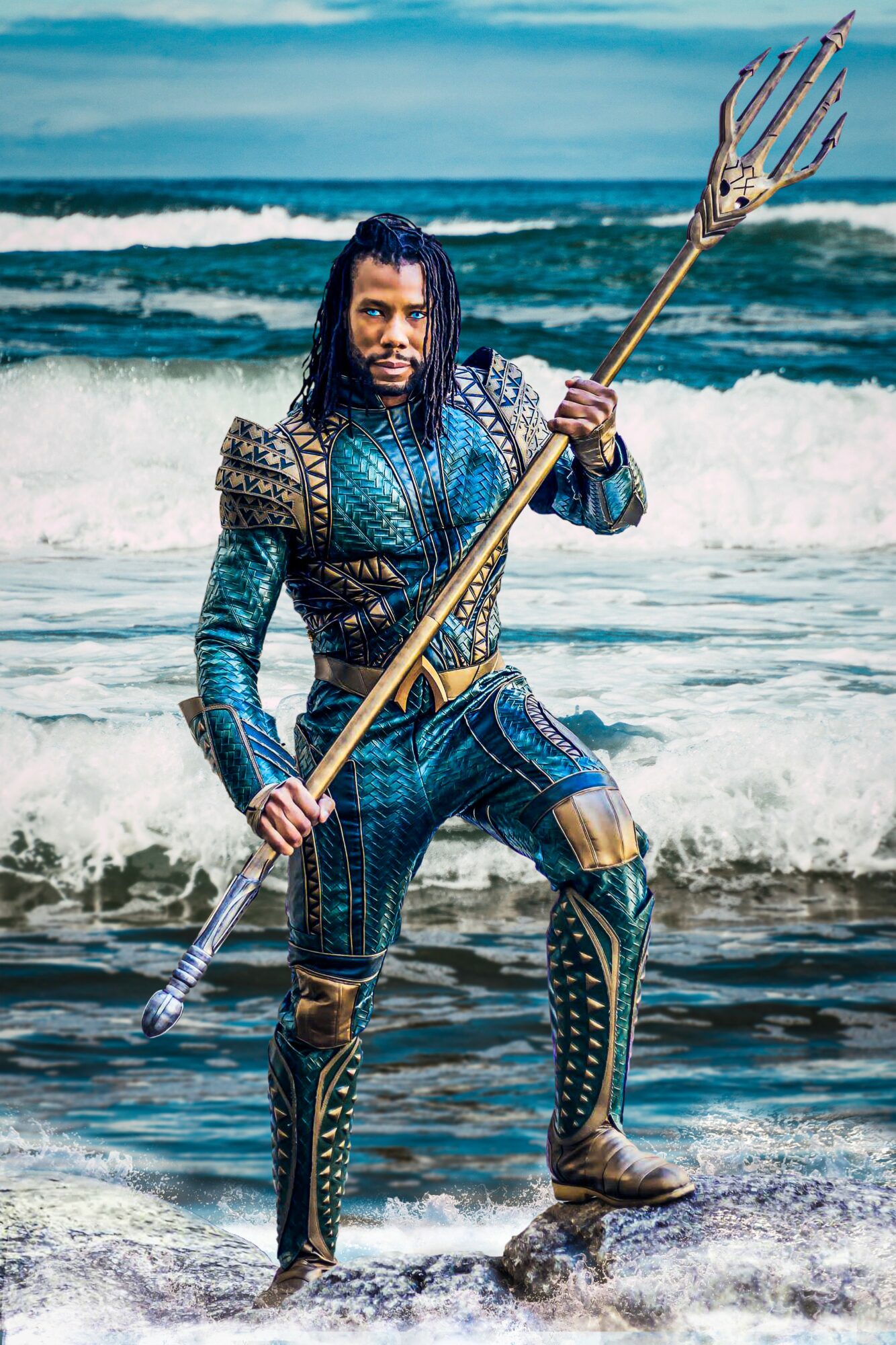 Person dressed as Aquaman holding a trident on the beach with ocean waves behind them.
