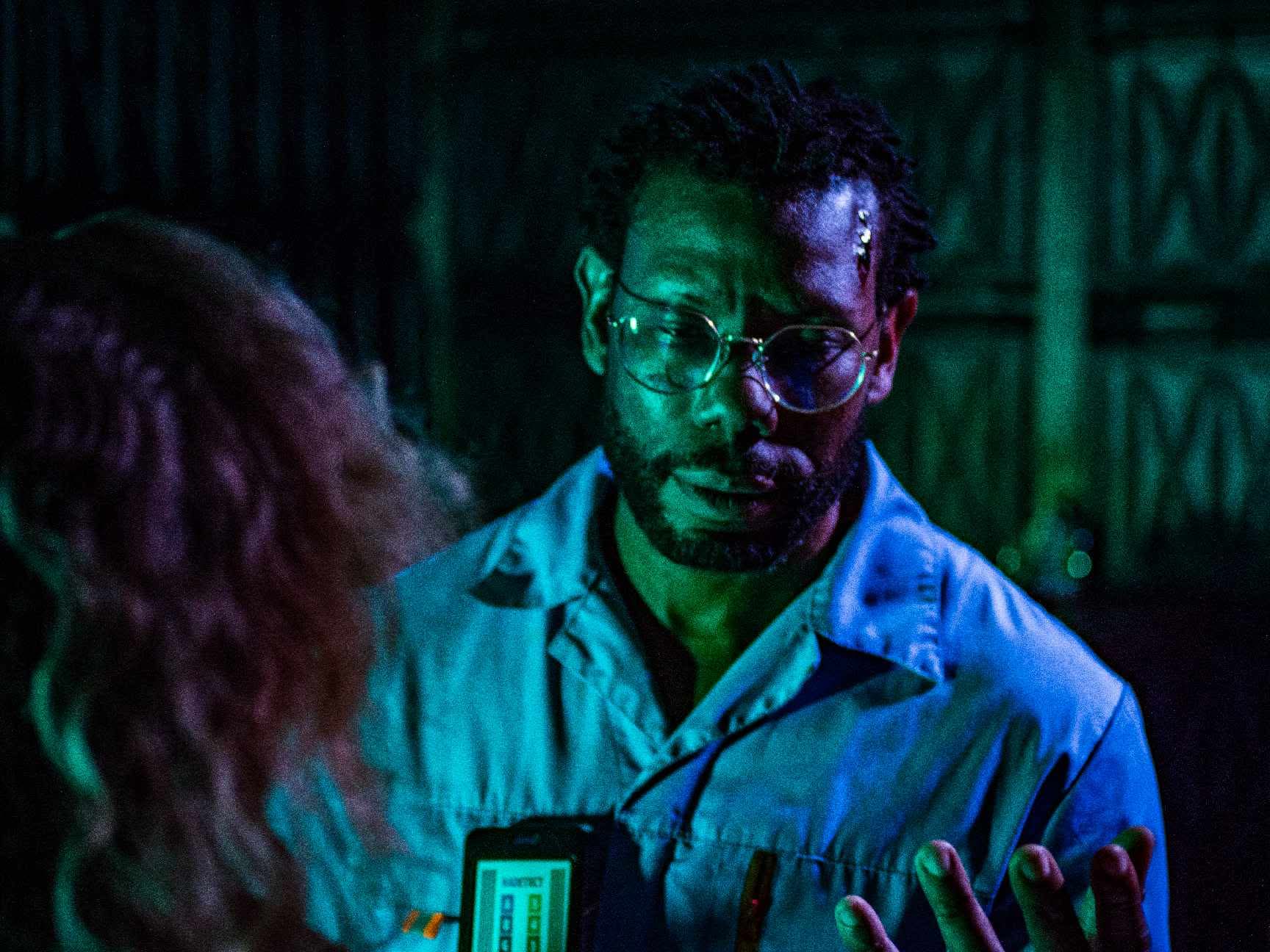 Man with glasses looking at a phone held by a person with curly hair, in a dimly lit room.
