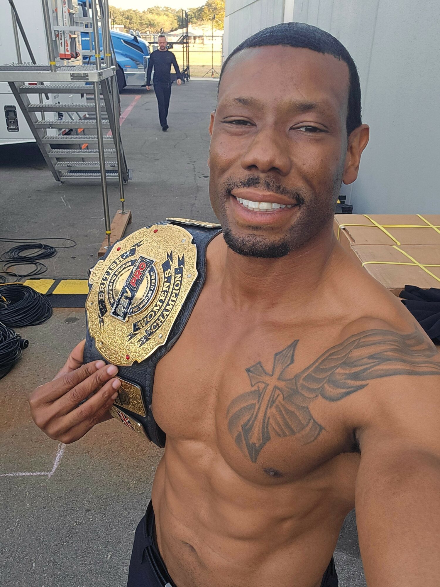 Man with a tattoo on his chest holding a championship belt, smiling, outdoors with a building in the background.