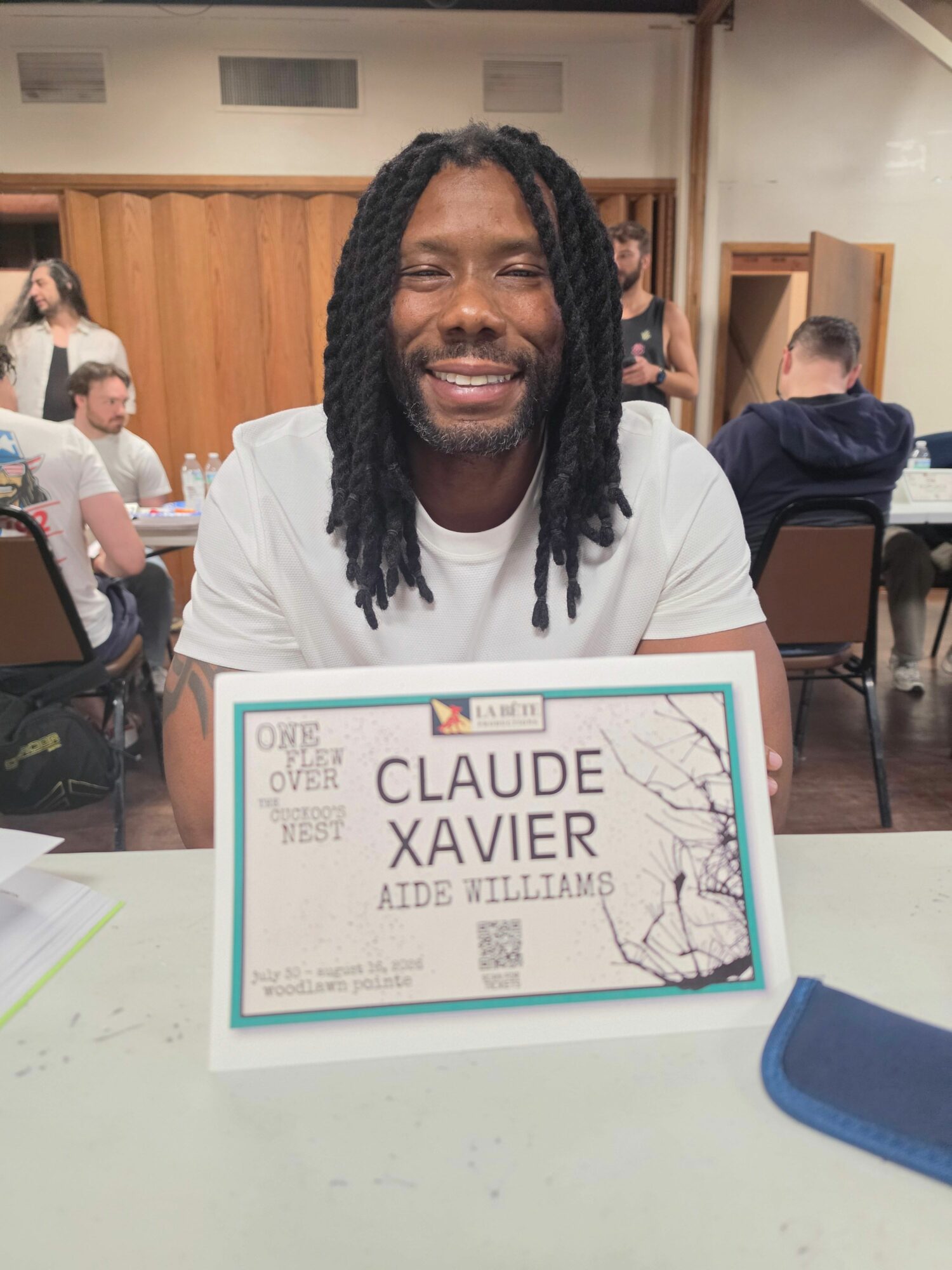 Person with long dreadlocks holding a certificate, smiling, in a room with other people and tables.