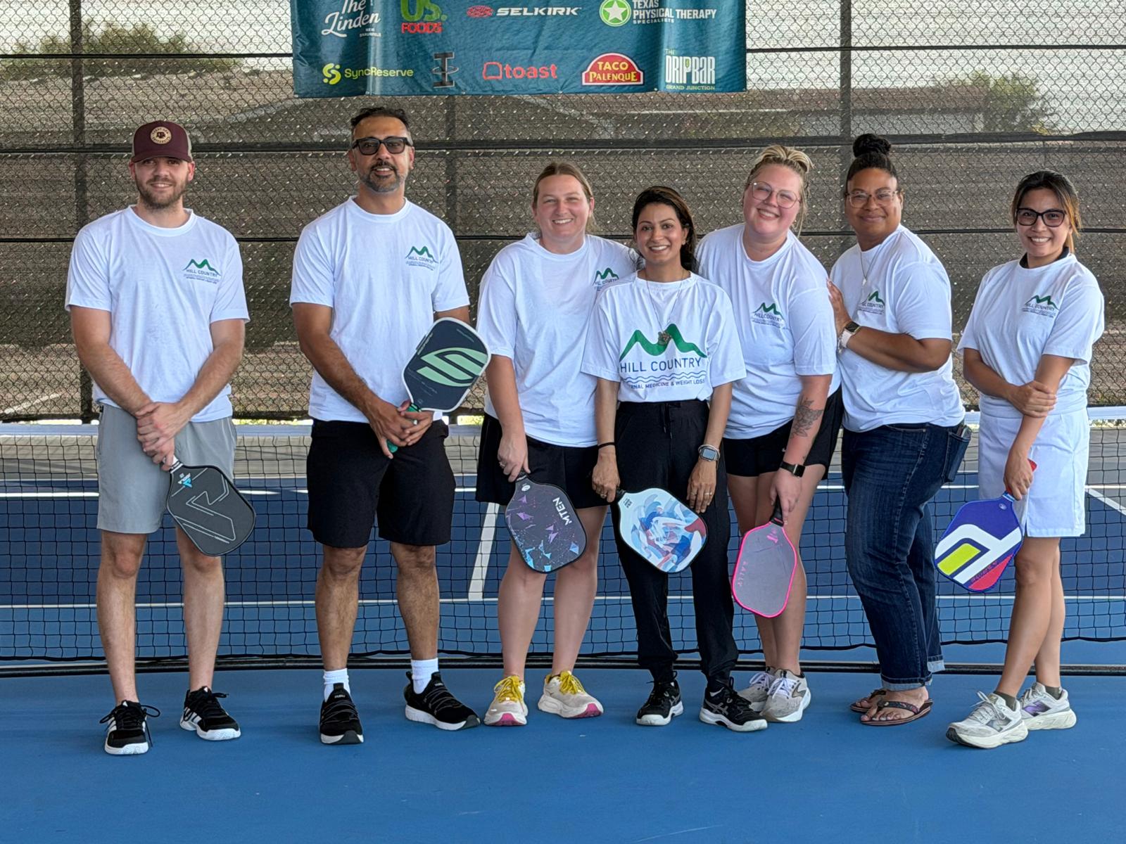 Group of seven people standing on a tennis court holding paddles, smiling, with a fence and banners in the background.