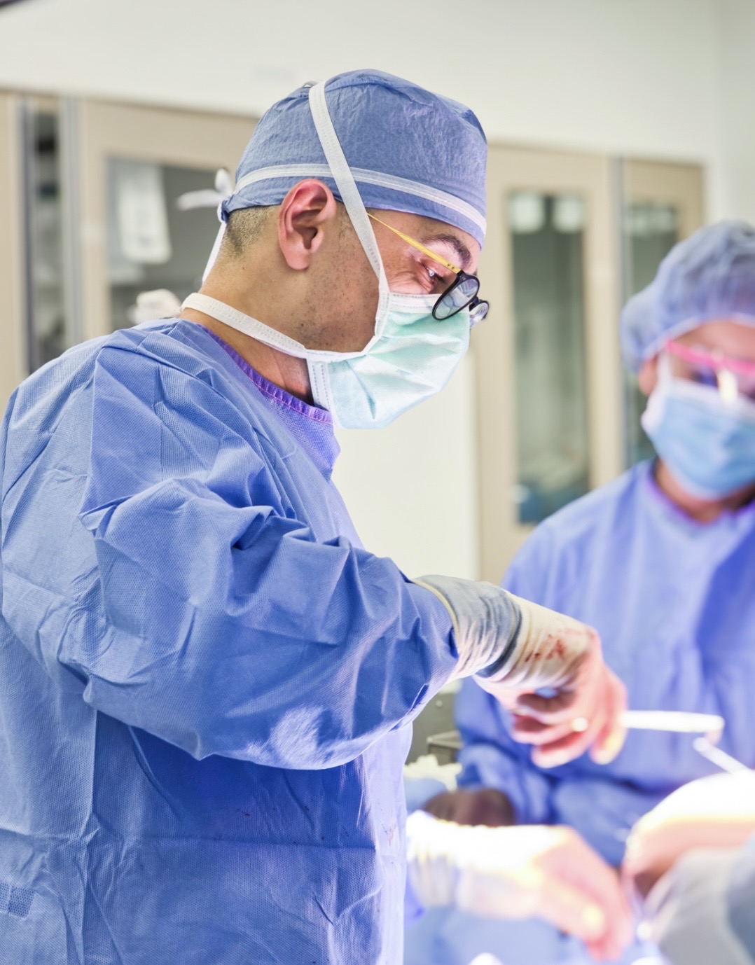 Surgeon in blue scrubs, mask, and glasses performs surgery with assistant in background, in operating room.